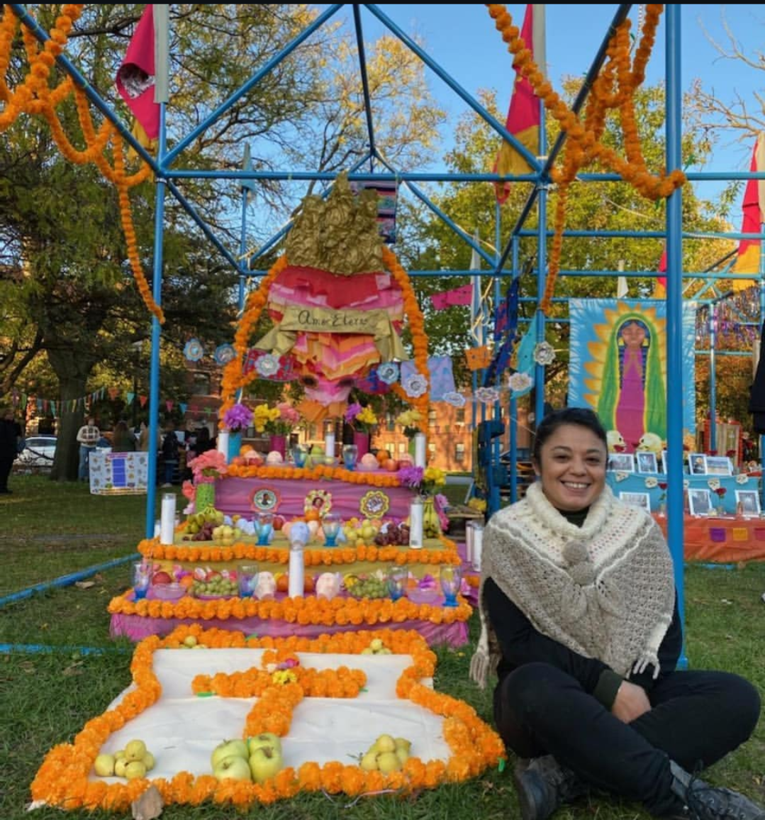 A woman sitting on the grass smiling in front of a colorful altar decorated with marigold flowers, fruits including apples, candles, and paper decorations, with a large religious icon or statue at the top of the altar. The scene is outdoors with trees and a blue sky in the background, indicating a festive celebration.