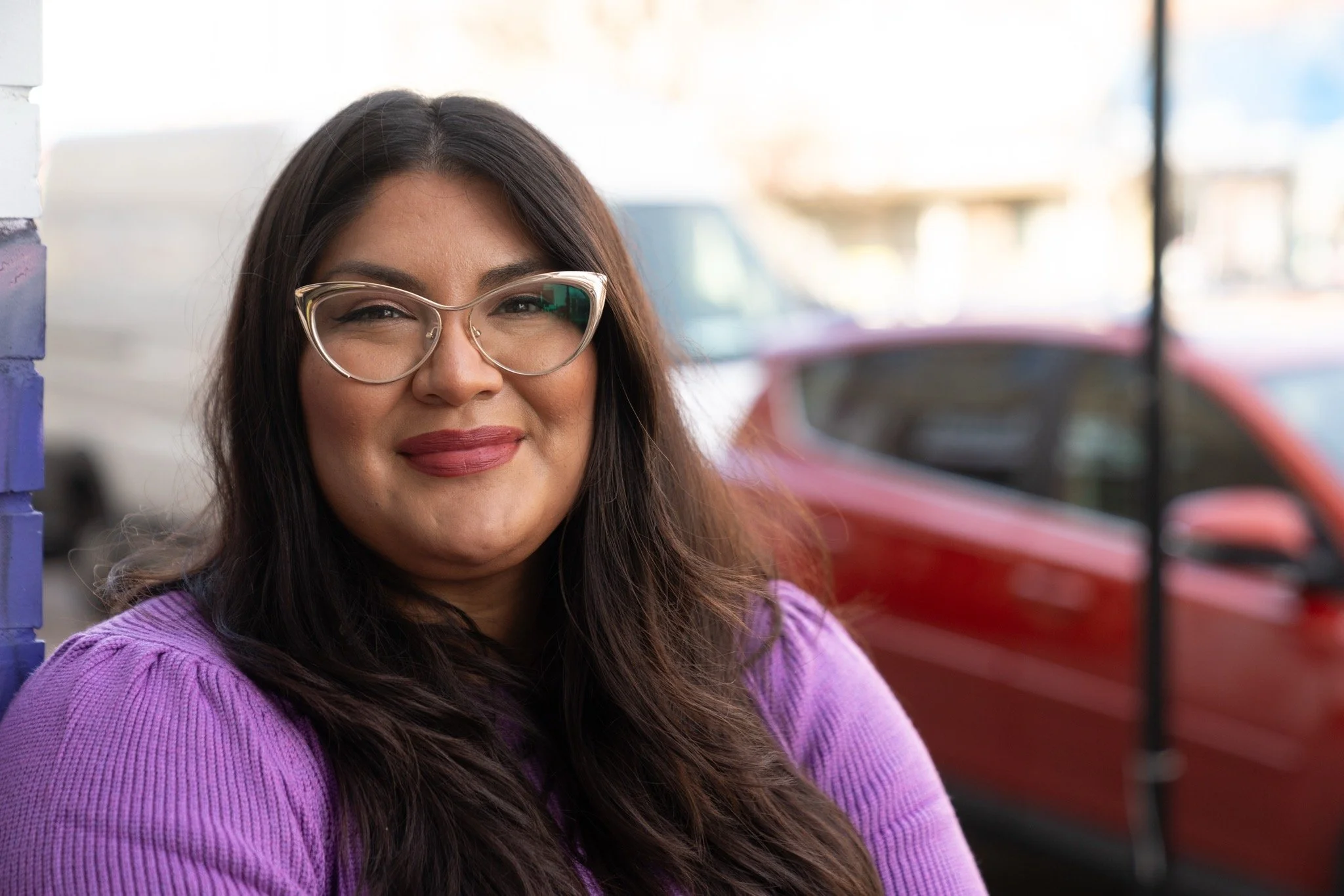 A woman with long dark hair wearing glasses and a purple sweater, smiling outdoors with a red car and a blurred background.
