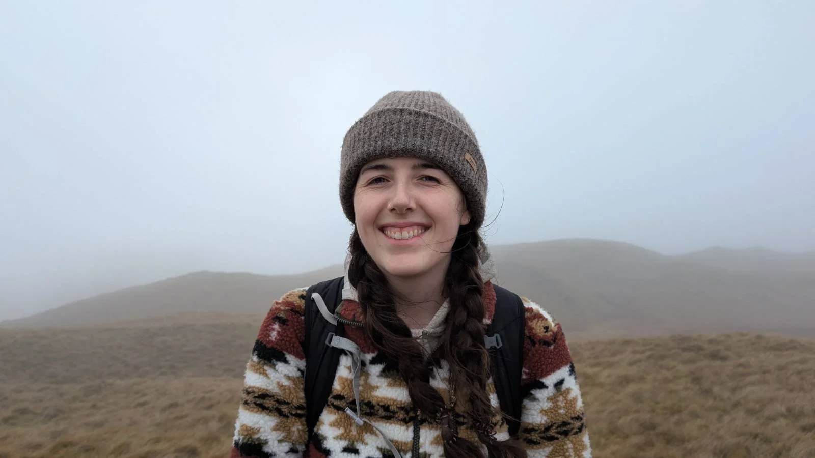 A woman with long braided hair, wearing a knit hat and patterned fleece jacket, smiling outdoors in a foggy hilly landscape.