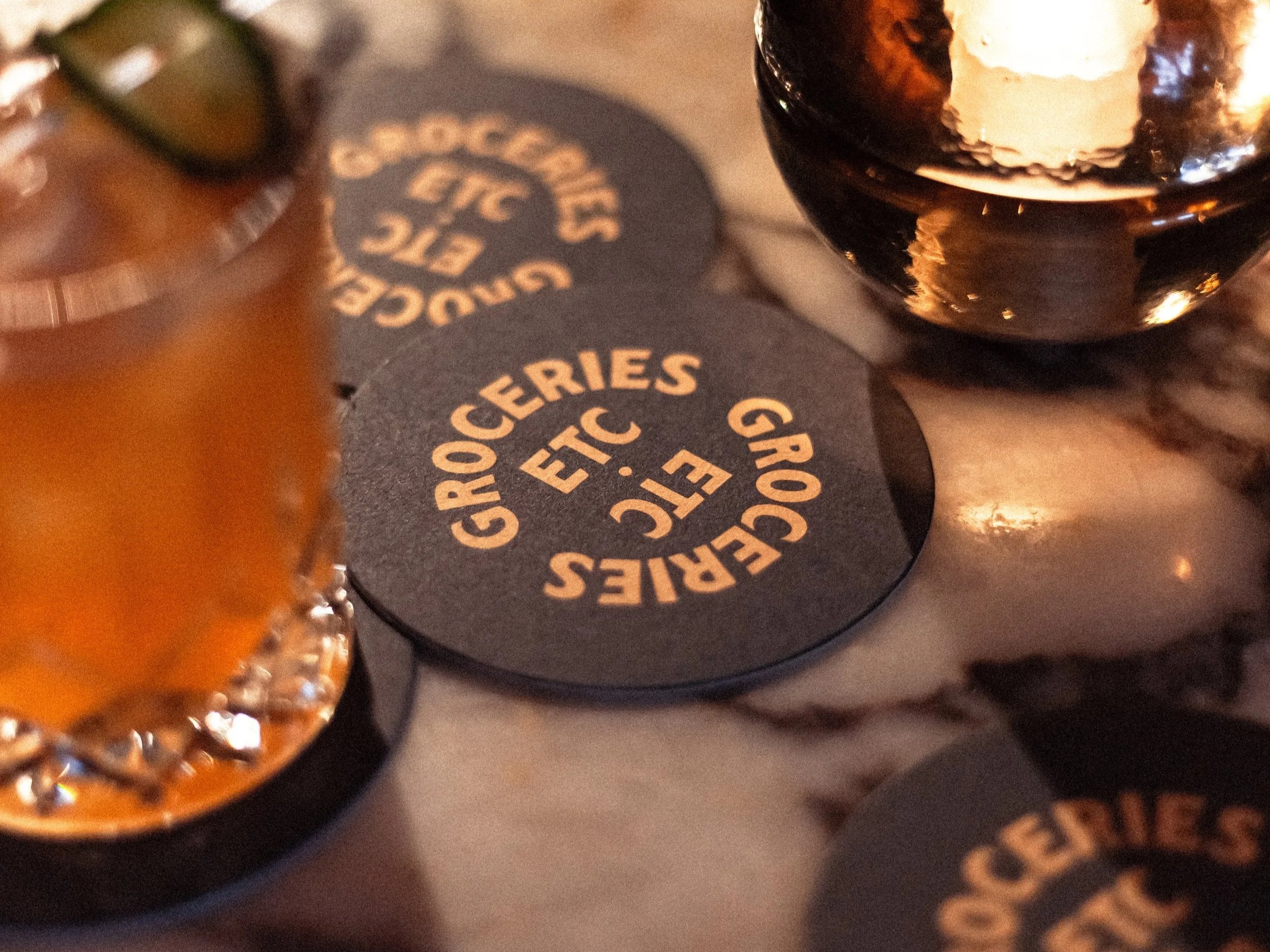 Close-up of a coaster with the text 'GROCERIES ETC.' and a glass of dark beverage with ice on a table.