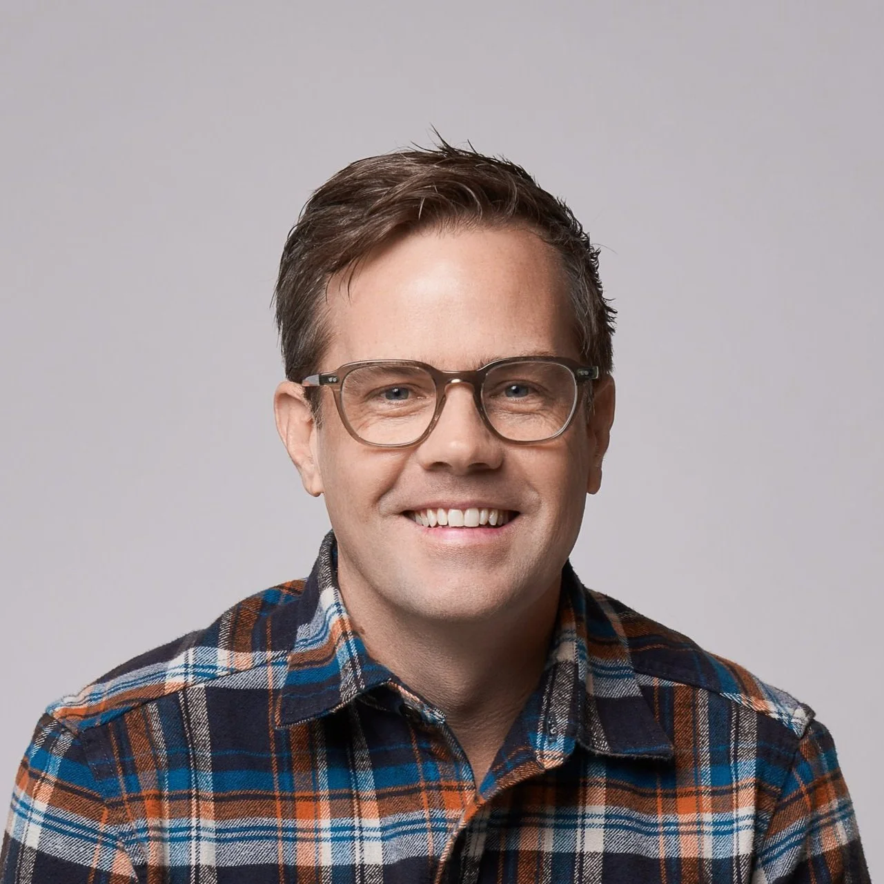 A smiling man with short brown hair and glasses, wearing a blue, brown, and white plaid shirt, against a plain gray background.