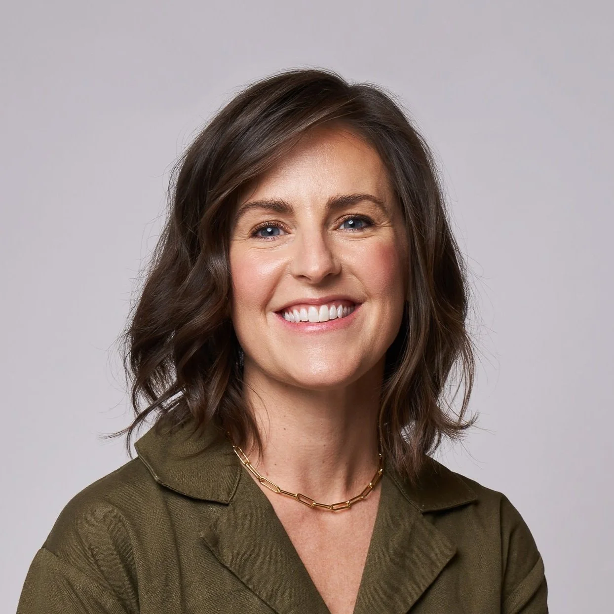 Headshot of a woman with shoulder-length wavy brown hair, blue eyes, smiling, wearing an olive green shirt and a gold chain necklace, against a plain light gray background.