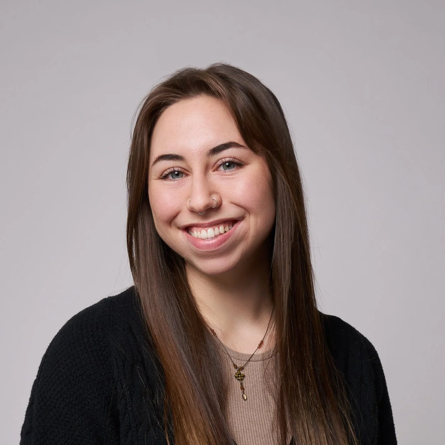 A young woman with long brown hair, light skin, and blue eyes, smiling and wearing a black cardigan and a gold necklace, against a plain gray background.
