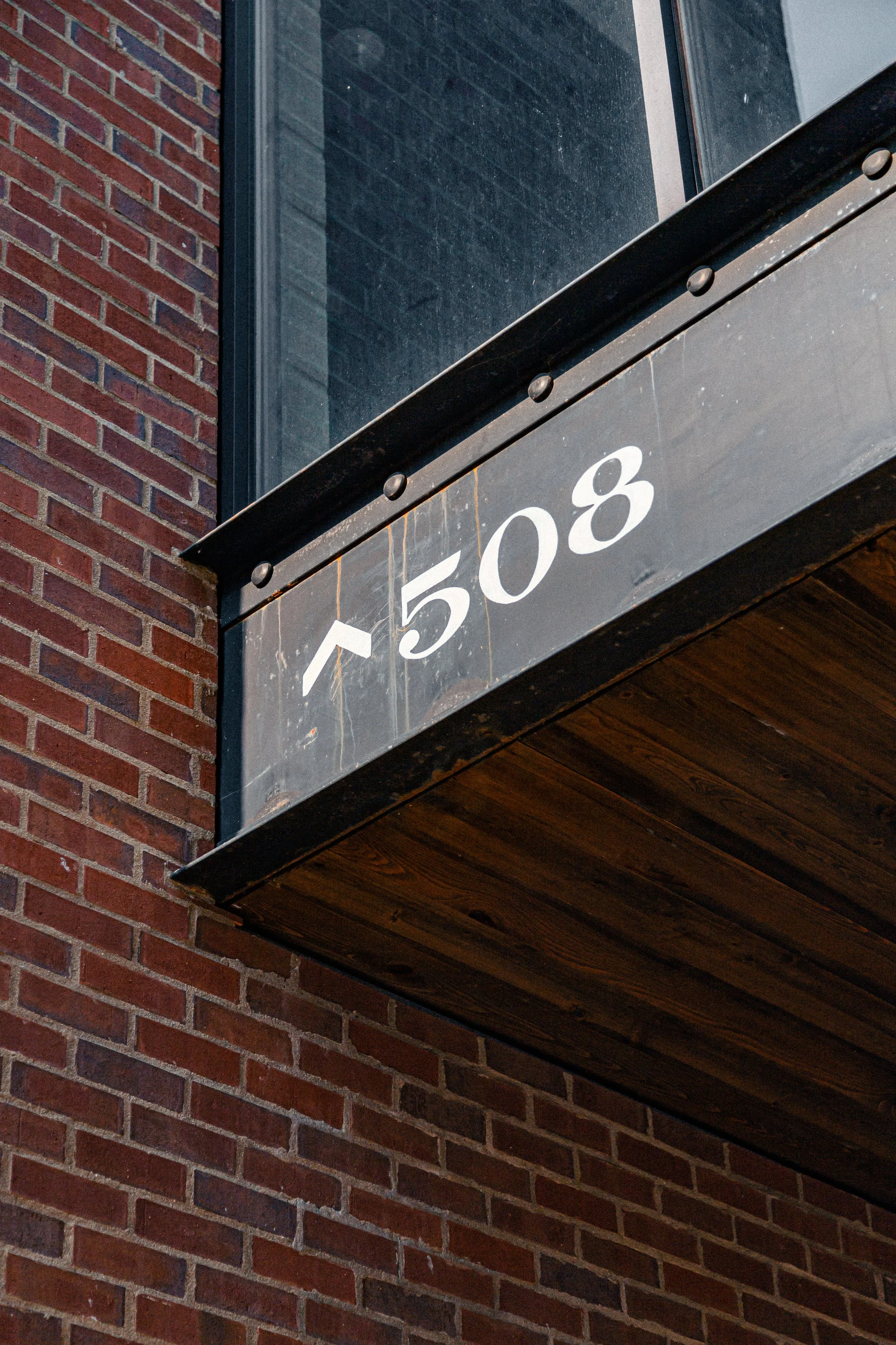 Close-up view of a building's exterior showing red brick wall on the left and a black rectangular sign with white numbers '5058' attached under a large window with black frame. The sign is mounted on a concrete ledge with a wooden underside.
