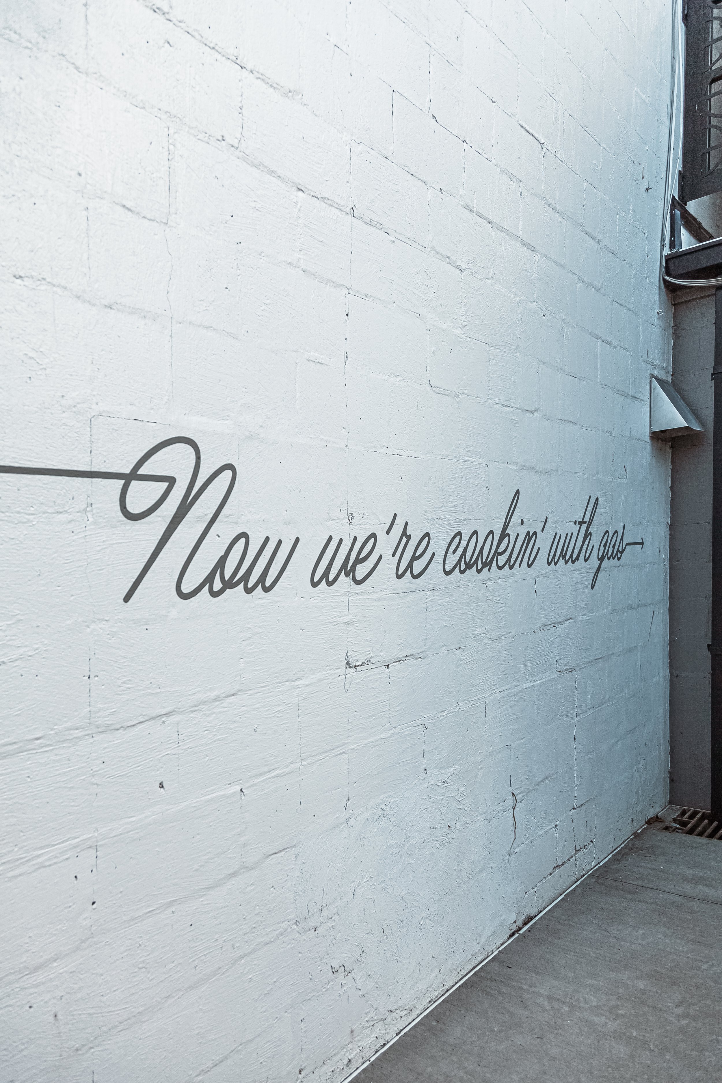 A white brick wall with black cursive writing that reads, 'Now we're cookin' with gas.' The wall is part of a room with a metal shelf and a floor drain visible.