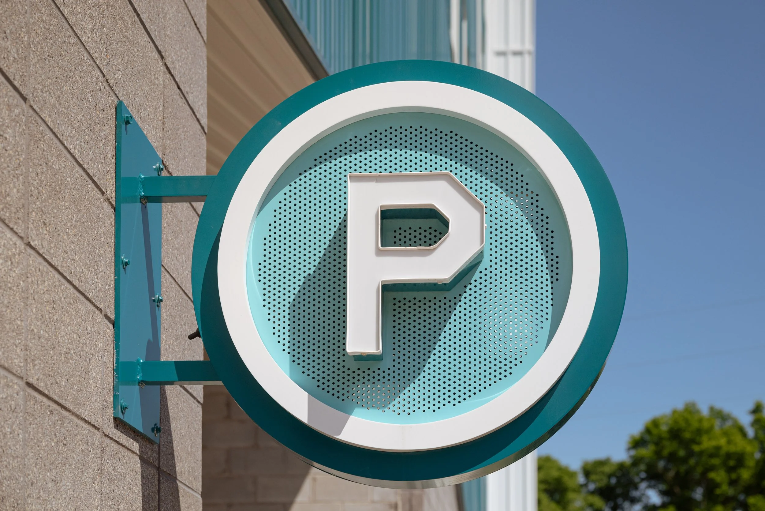 A teal sign with a white letter P for parking, mounted on a brick wall outdoors, with a blue sky in the background.