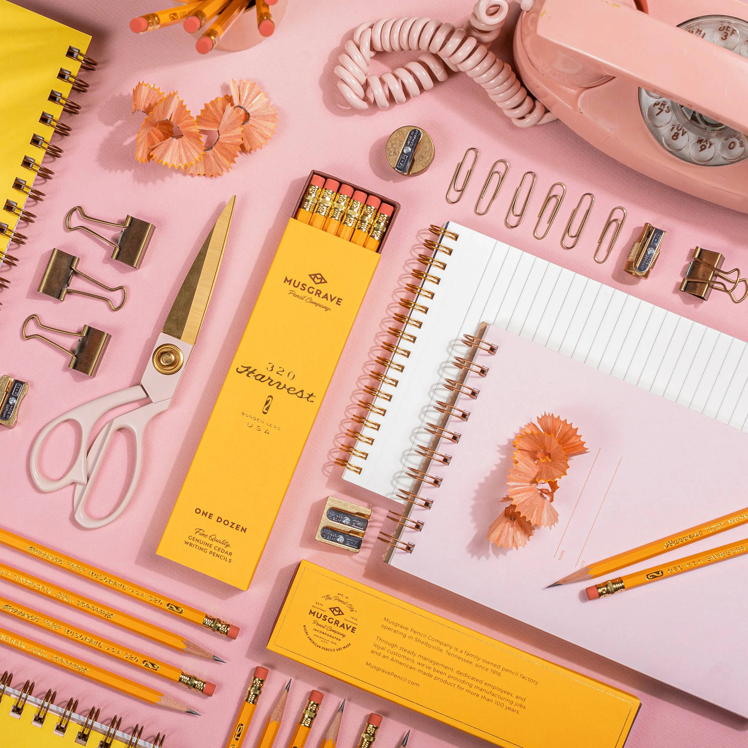 Office desk setup with yellow notebooks, pink pencils, pink rotary phone, scissors, paper clips, orange pencil shavings, and a small notepad on a pink background.