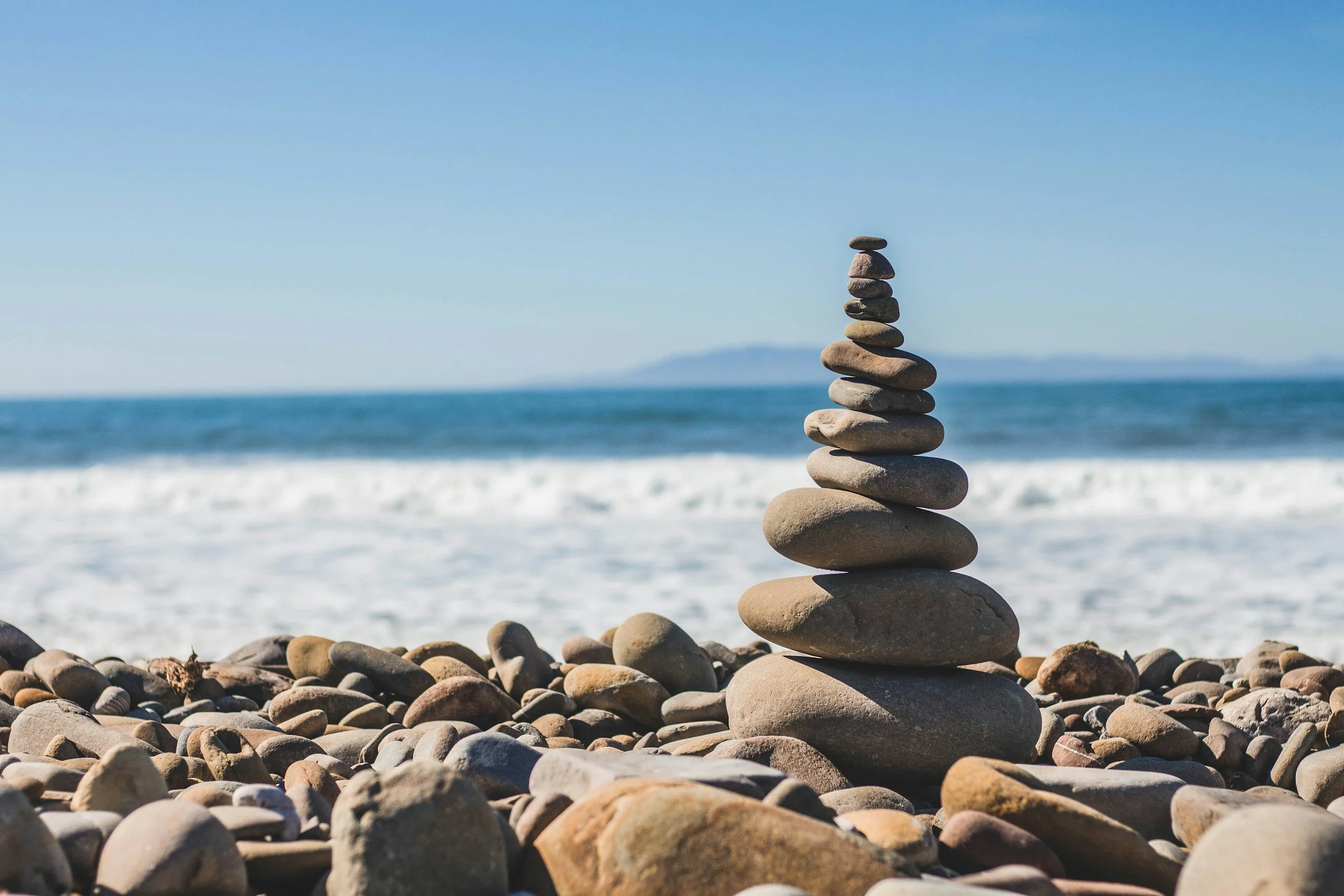A stack of smooth rounded stones on a beach, with the ocean and distant land in the background under a clear blue sky.