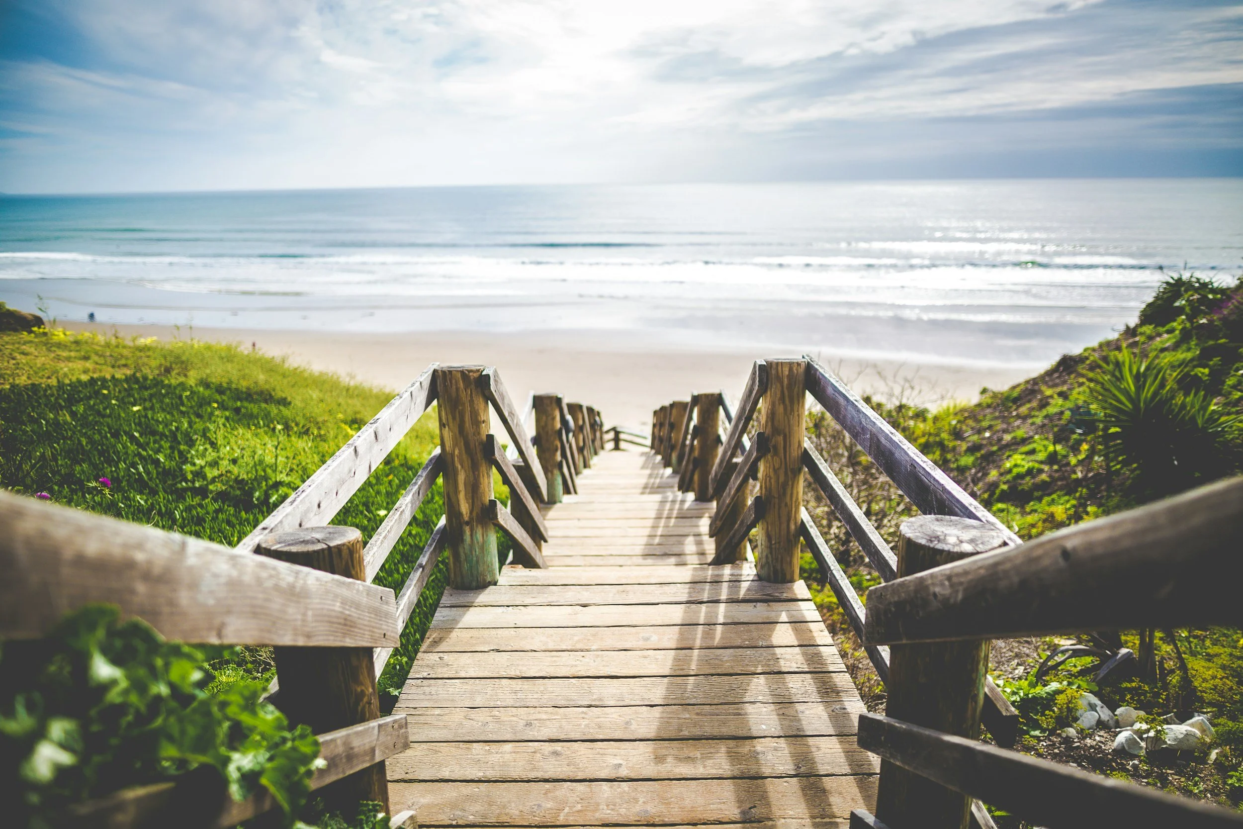 Wooden stairs leading down to a sandy beach with the ocean in the background and a cloudy sky.