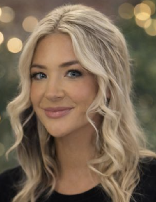 A woman with long, wavy blonde hair and blue eyes smiling at the camera, with a blurred background of warm lights.