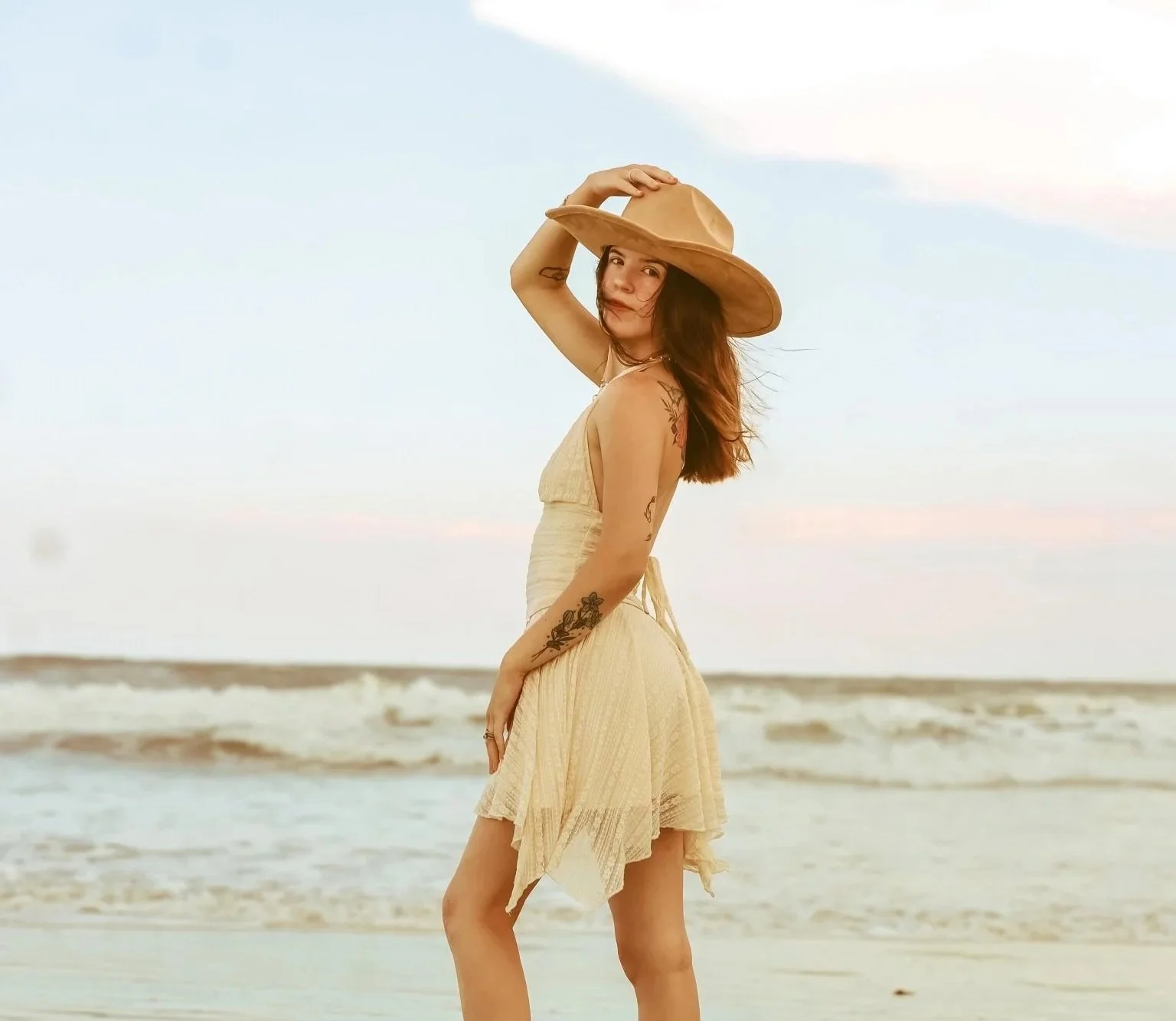A woman with tattoos in a light dress and wide-brimmed hat standing on a sandy beach with ocean waves and a pastel sky in the background.