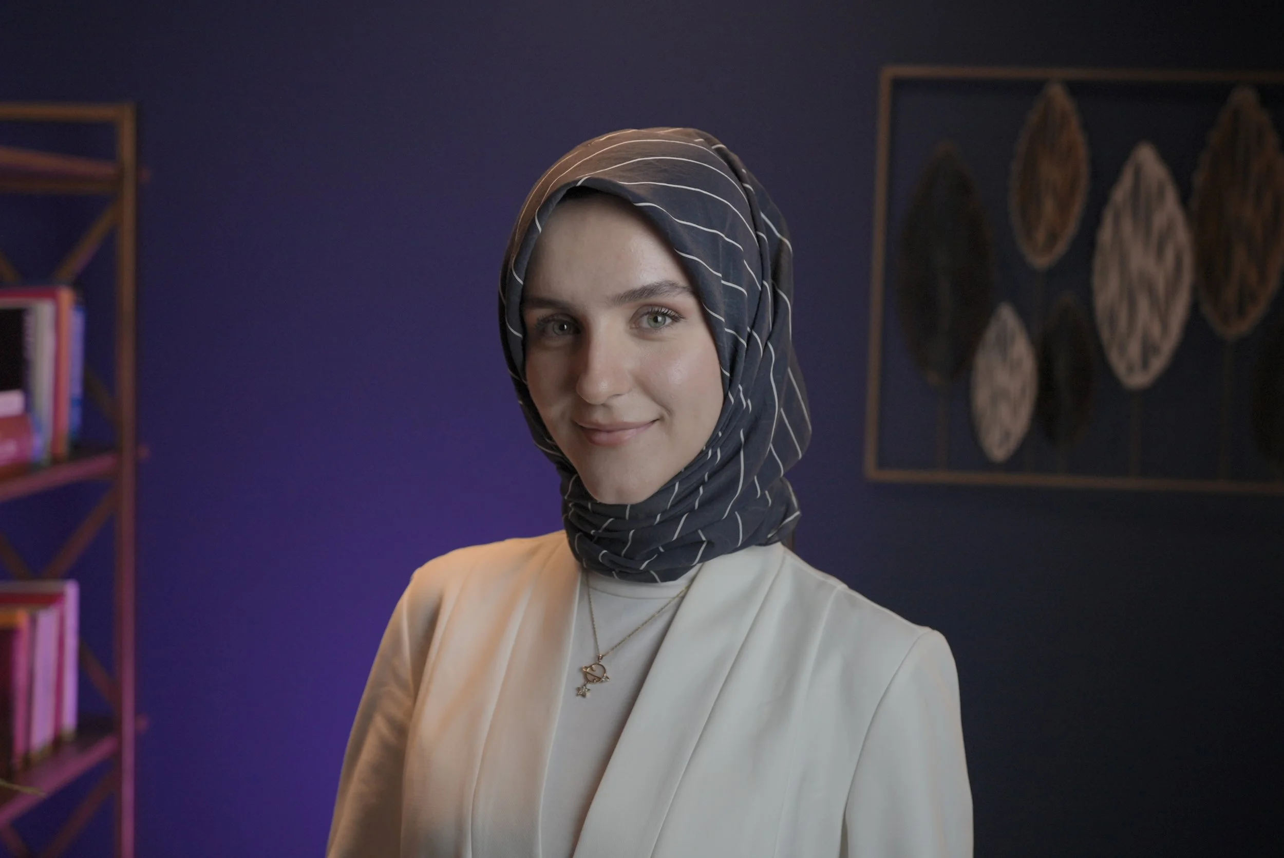 A woman wearing a dark striped headscarf and a white top, standing in a room with a purple wall and framed artwork in the background.