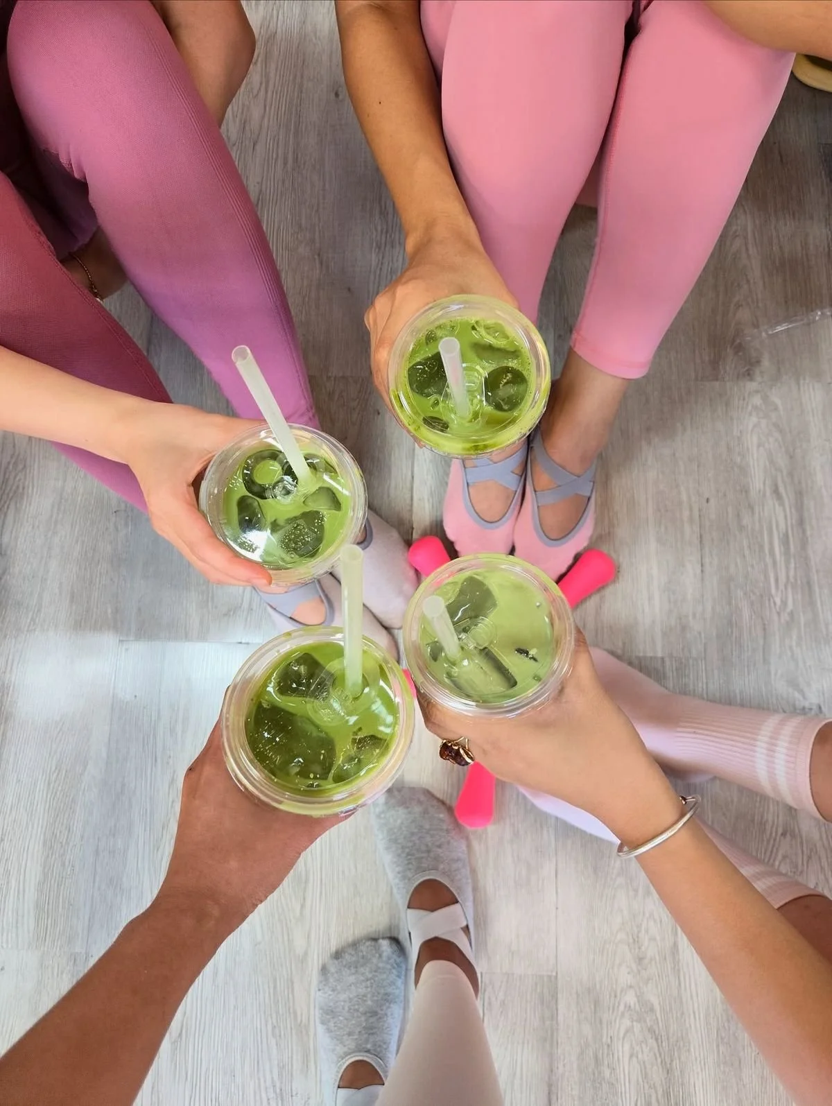 Four people are holding cups of green iced beverages with ice cubes and straws, seen from above, while sitting on a wooden floor. They are wearing pastel-colored pants and pastel socks or slippers.