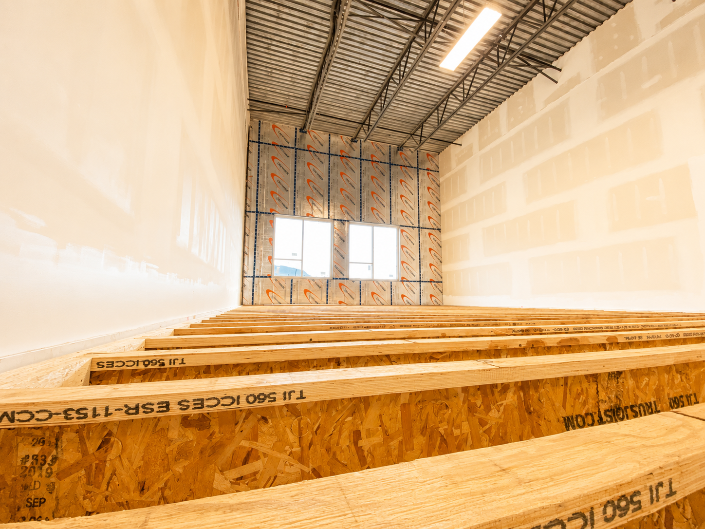 An unfinished room under construction with exposed wall studs, uninsulated walls, and two windows on one wall. The ceiling has visible metal framing and a fluorescent light fixture.