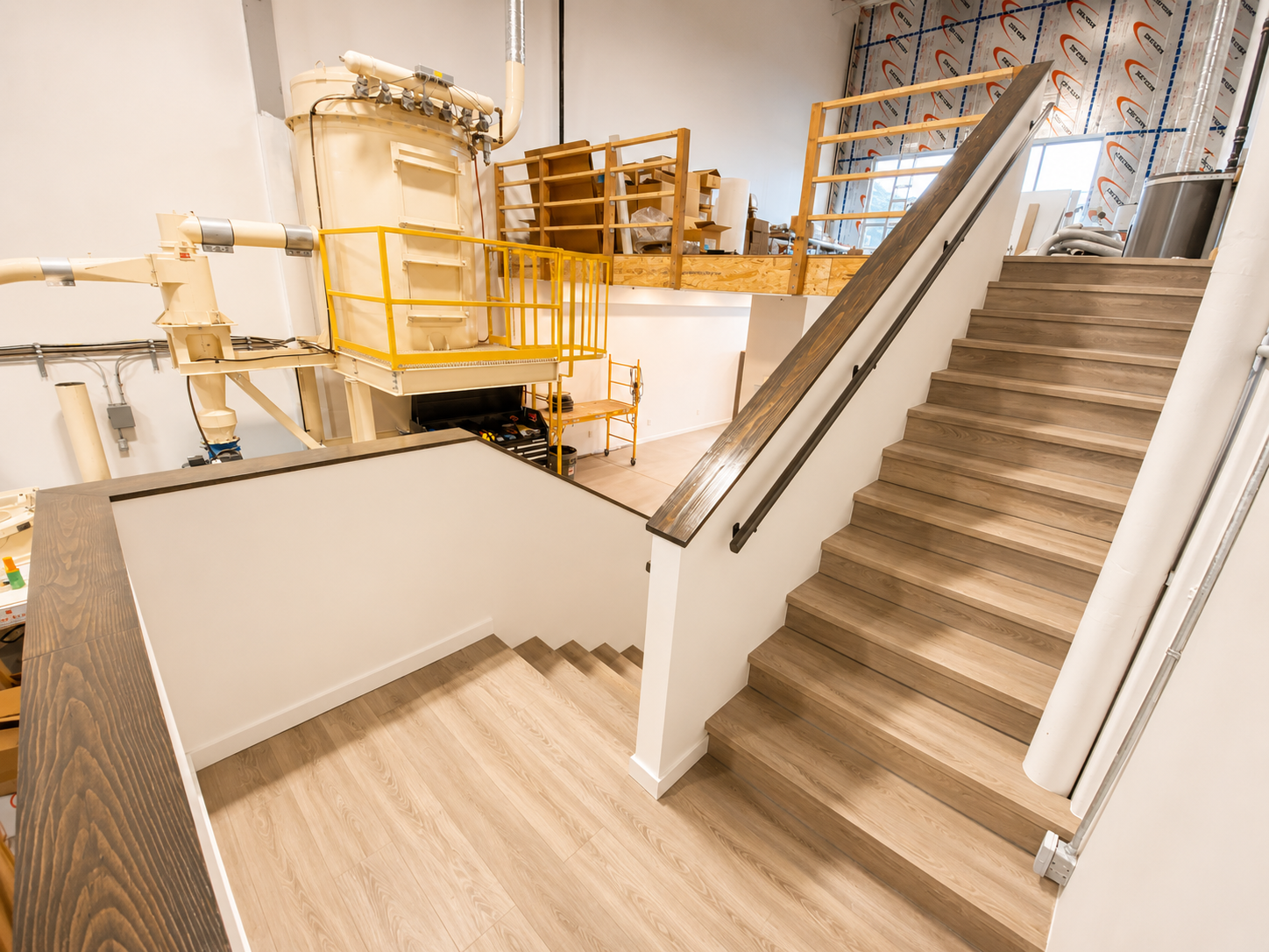 Interior view of a building under construction showing a staircase with wooden steps, a safety railing, and industrial equipment in the background.