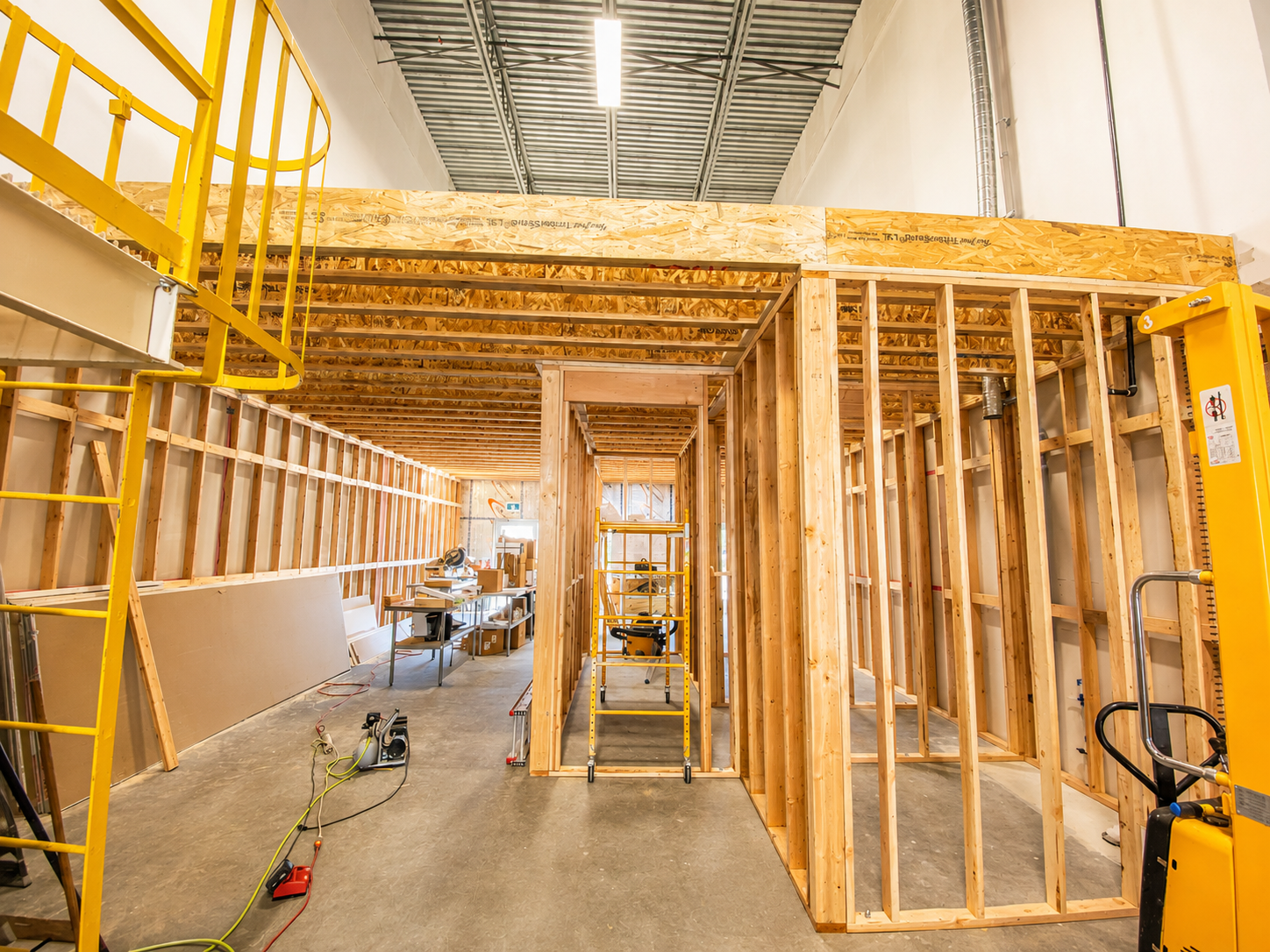Interior view of a construction site with wooden framing for walls and ceiling, construction tools and equipment, scaffolding, and natural light from a window.
