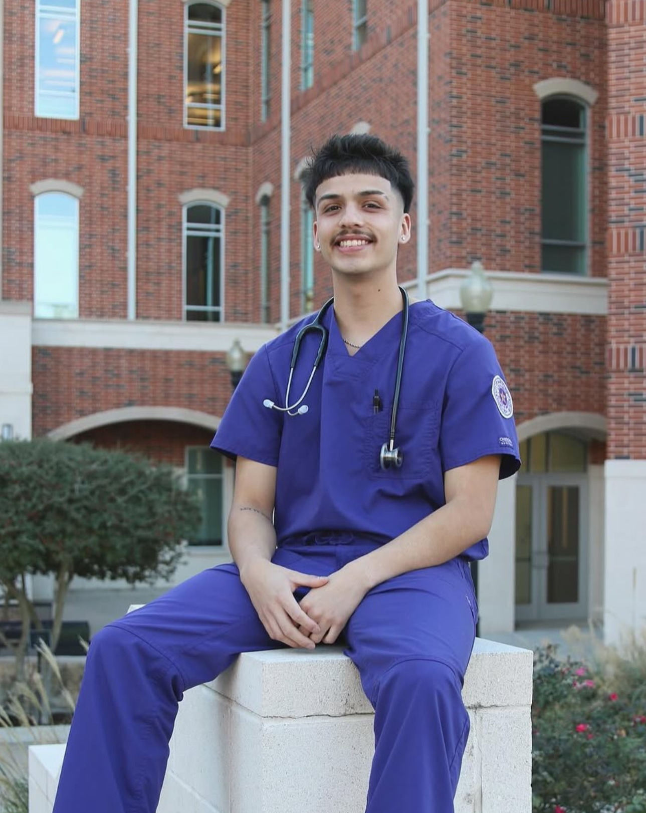 Young man wearing blue scrubs and a stethoscope sitting outdoors in front of a brick building.