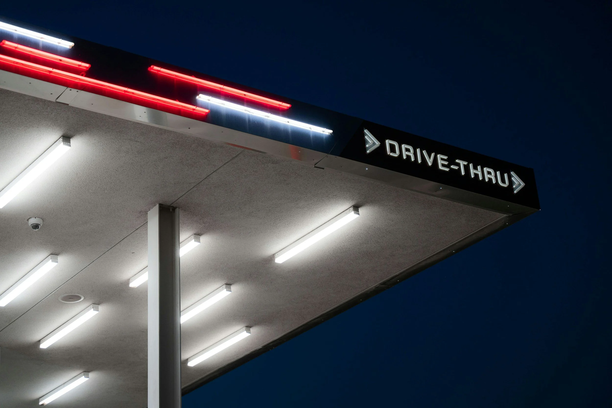 Night view of a drive-thru sign with illuminated arrow and text, under a dark sky, showing a modern building with lit ceiling lights.