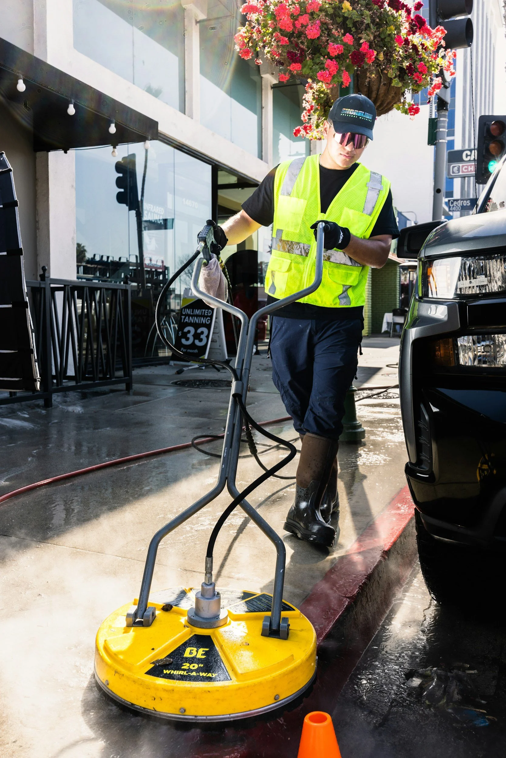 A person in a black t-shirt, yellow safety vest, and rubber boots is cleaning the sidewalk with a ride-on floor buffer. The person is wearing sunglasses and a cap. There is a black vehicle parked nearby, a hanging basket of pink and red flowers above, and a storefront with large glass windows in the background.