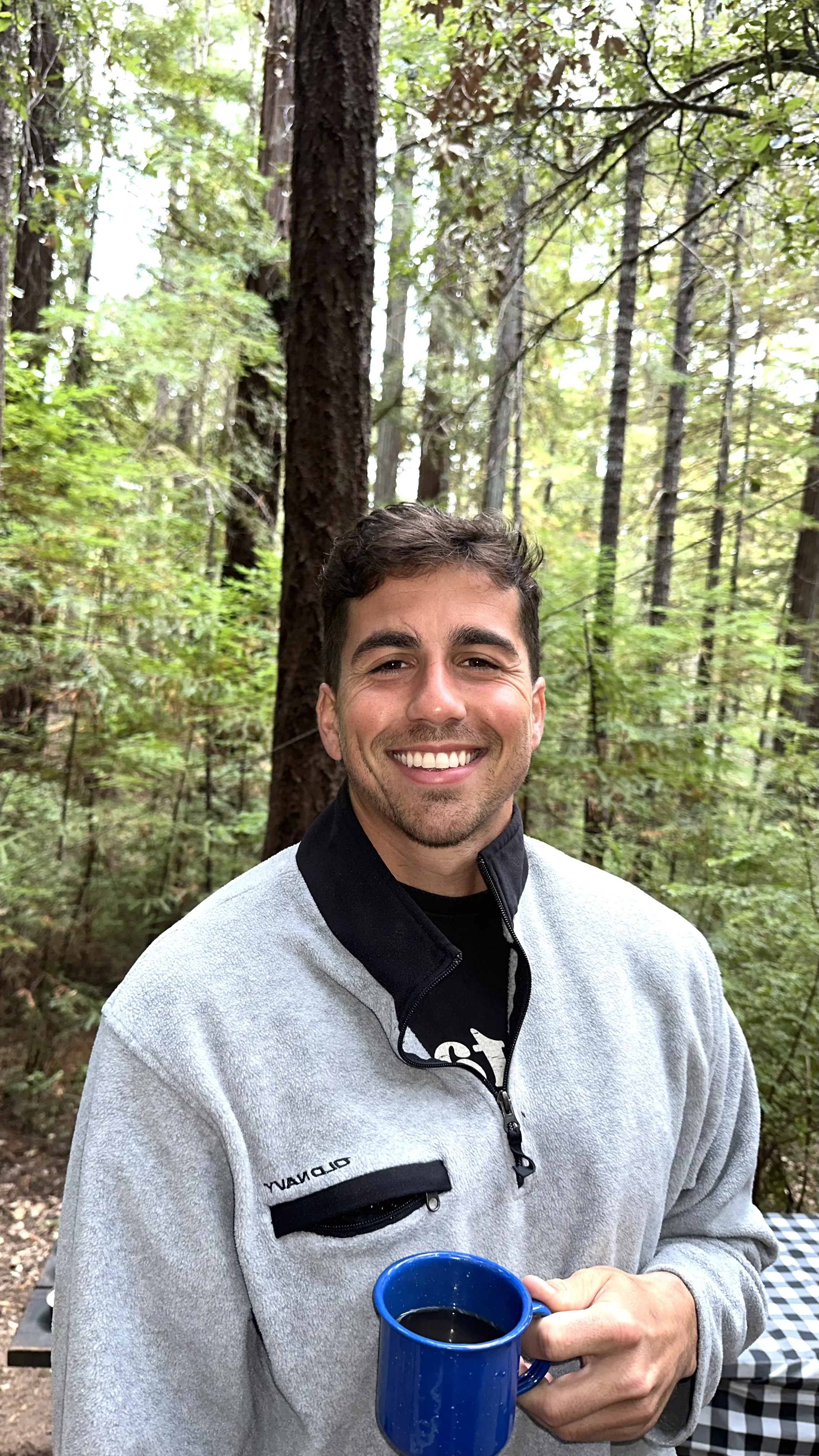 A young man smiling and holding a blue mug of coffee outdoors in a forest setting with tall trees and green foliage.