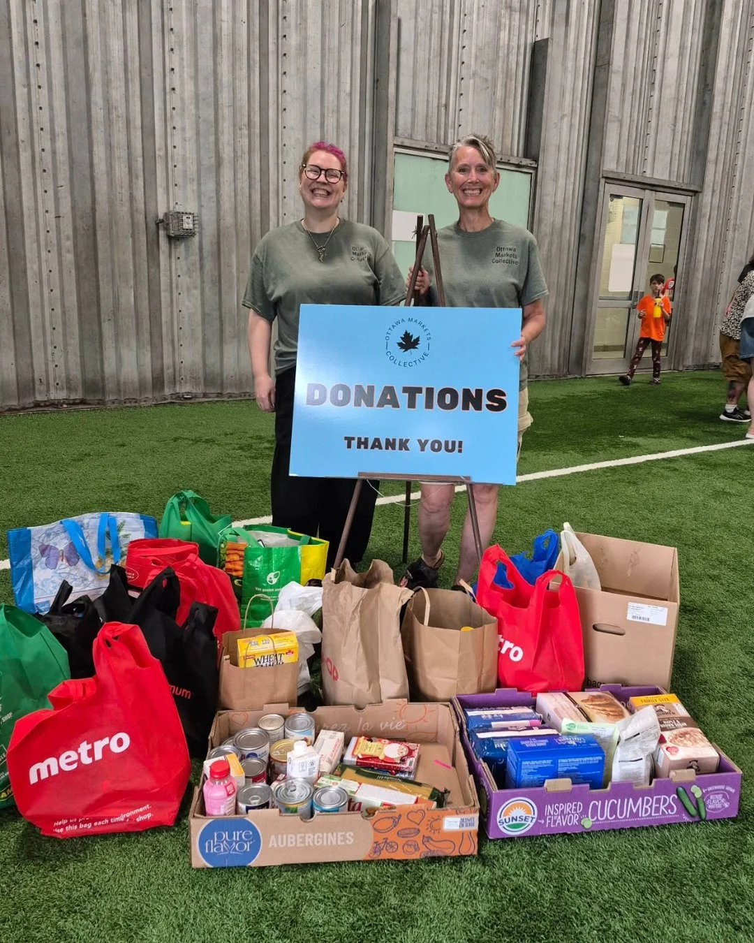 Two women standing behind donated food and supplies, holding a sign that reads 'Donations Thank You!' in an indoor sports facility with a green turf floo.