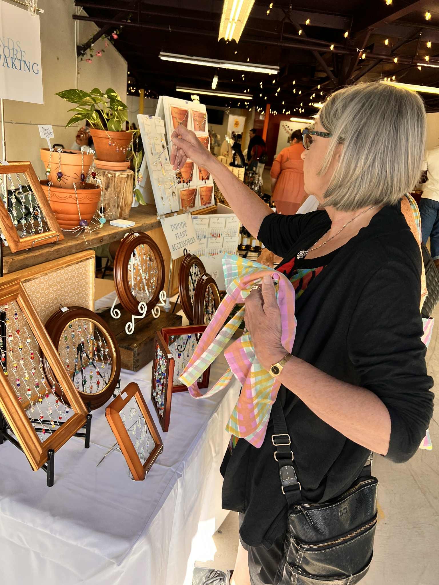 A woman with short blonde hair and glasses is shopping at a jewelry display table, examining various items at an indoor market.
