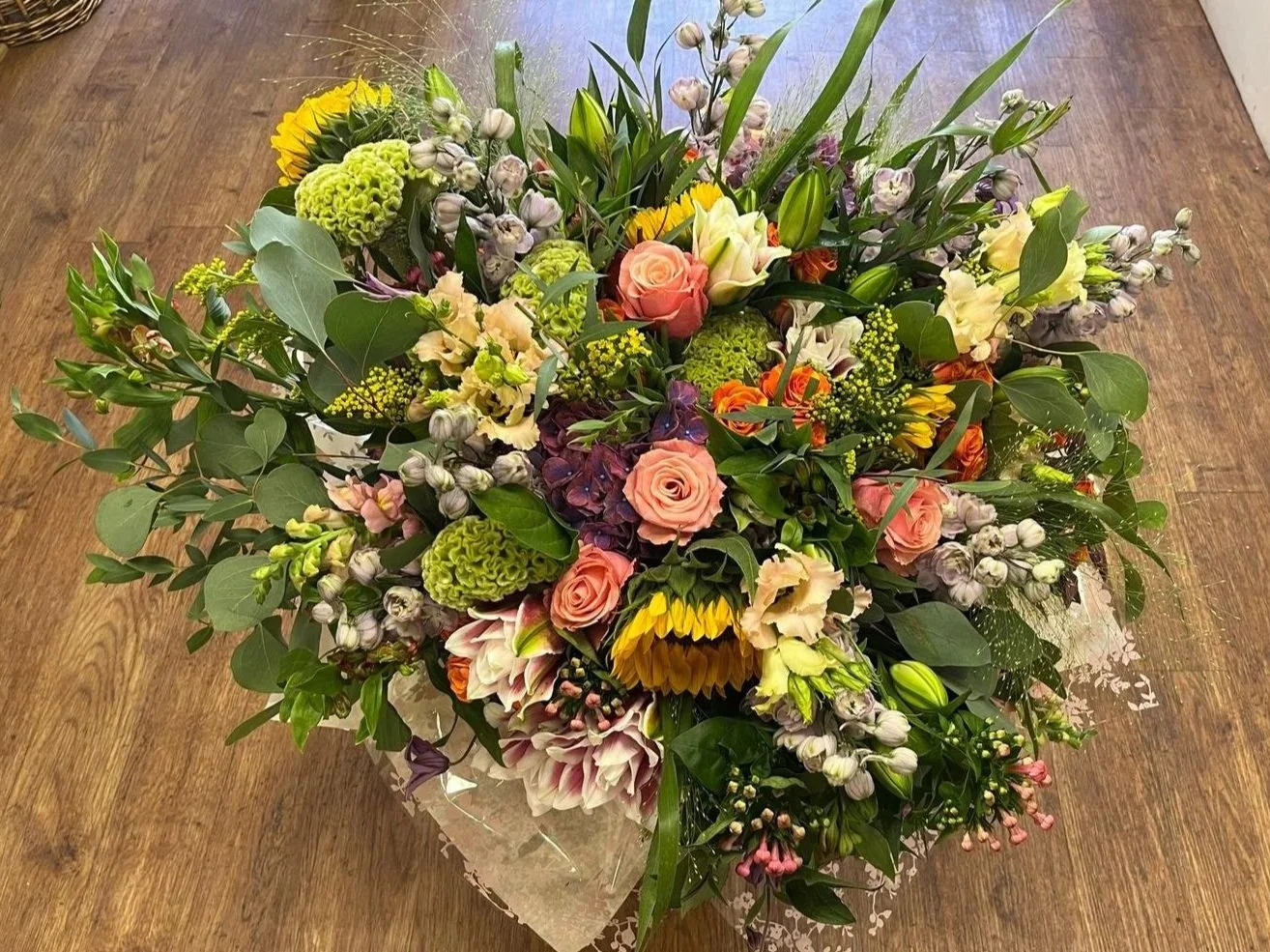 A colorful bouquet of various flowers, including roses, sunflowers, lilies, and greenery, arranged on a wooden table.