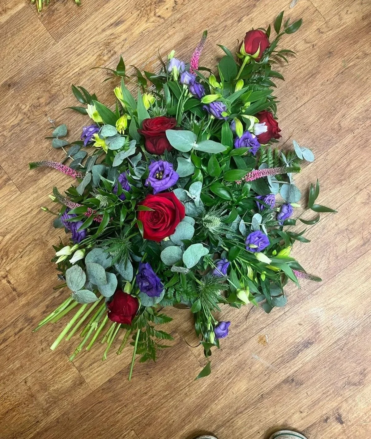 A colorful floral arrangement with red roses, purple lisianthus, white lilies, and greenery, arranged on a wooden floor.
