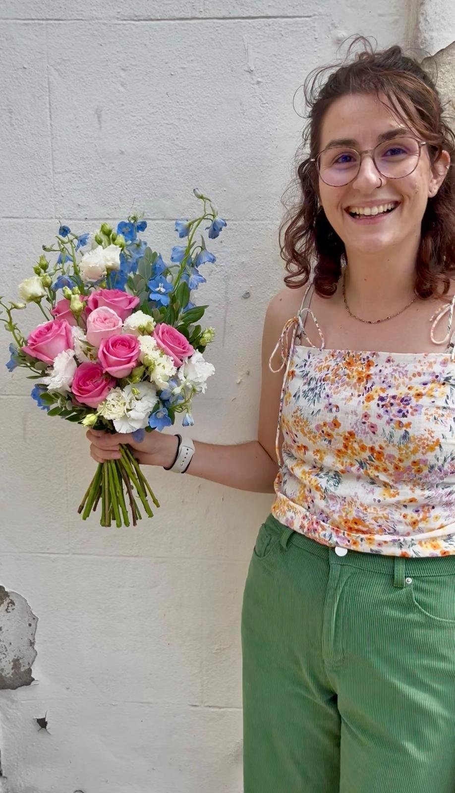 A young woman with glasses, wavy brown hair, and a tattoo on her arm, smiling and holding a colorful flower bouquet, standing against a white brick wall.