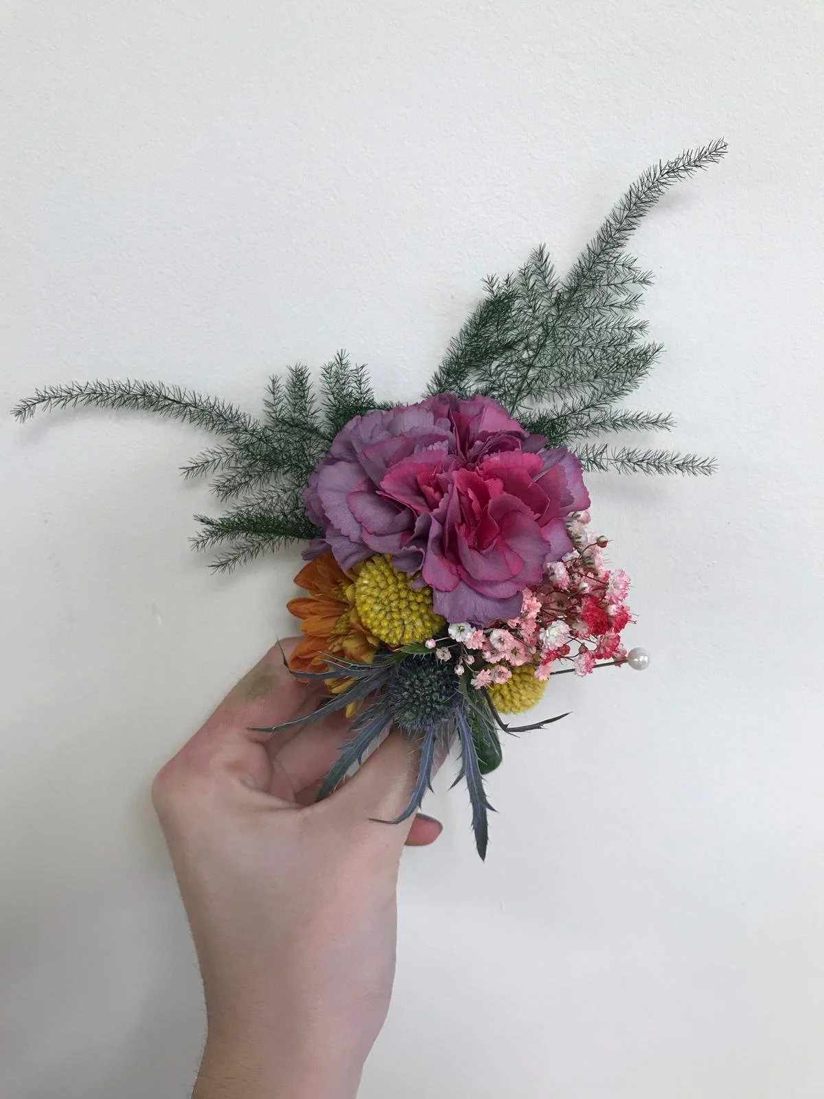Hand holding a small bouquet of mixed flowers with pink, purple, yellow, orange, and white blooms, accented with green foliage and a pearl pin, against a plain white background.