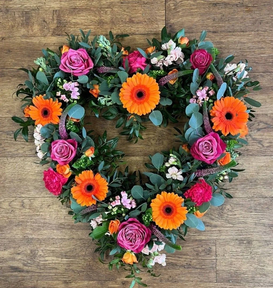 A heart-shaped floral arrangement on a wooden surface, featuring orange gerbera daisies, pink roses, and various green foliage.
