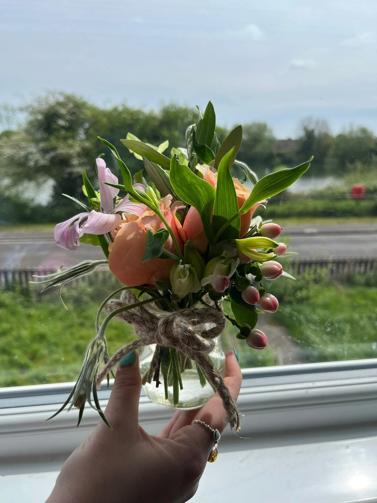 Person holding a small glass jar of a flower bouquet with pink lilies, peach roses, and green leaves against a window with a view of trees and sky outside.