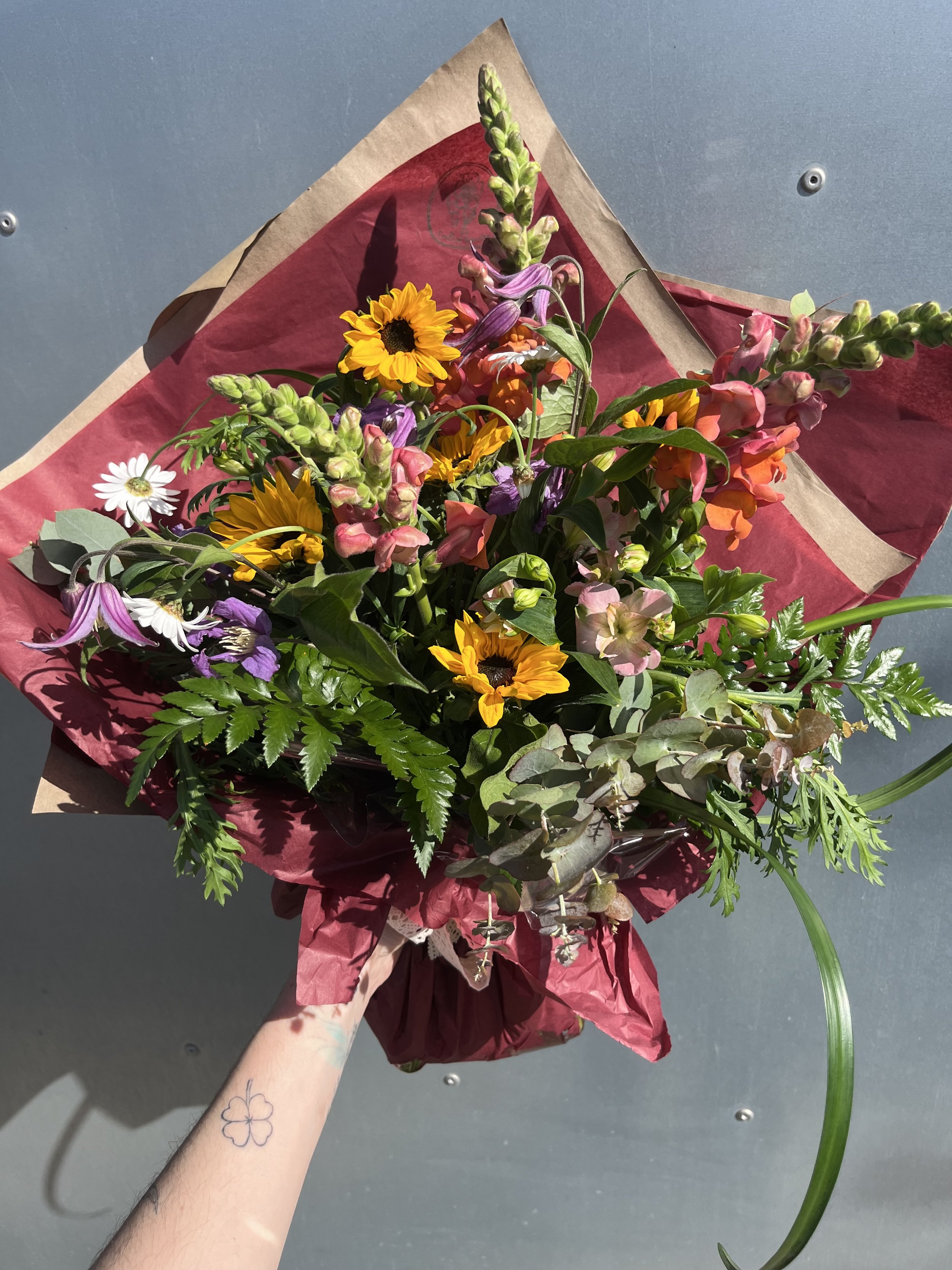 A hand holding a bouquet of colorful flowers wrapped in red and brown paper, set against a gray background.