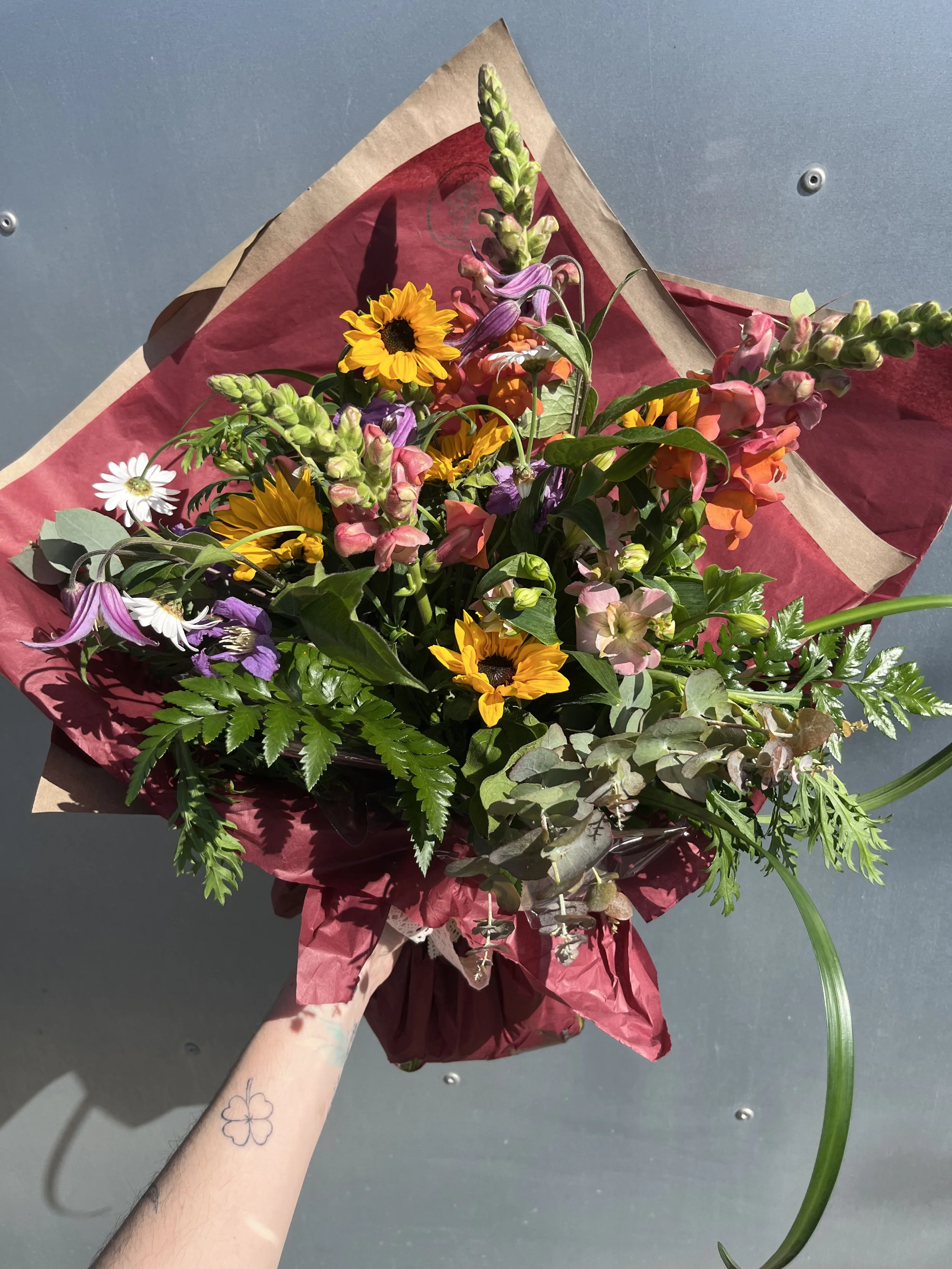 Colourful hand-tied bouquet with sunflowers, snapdragons and mixed flowers wrapped in burgundy paper.