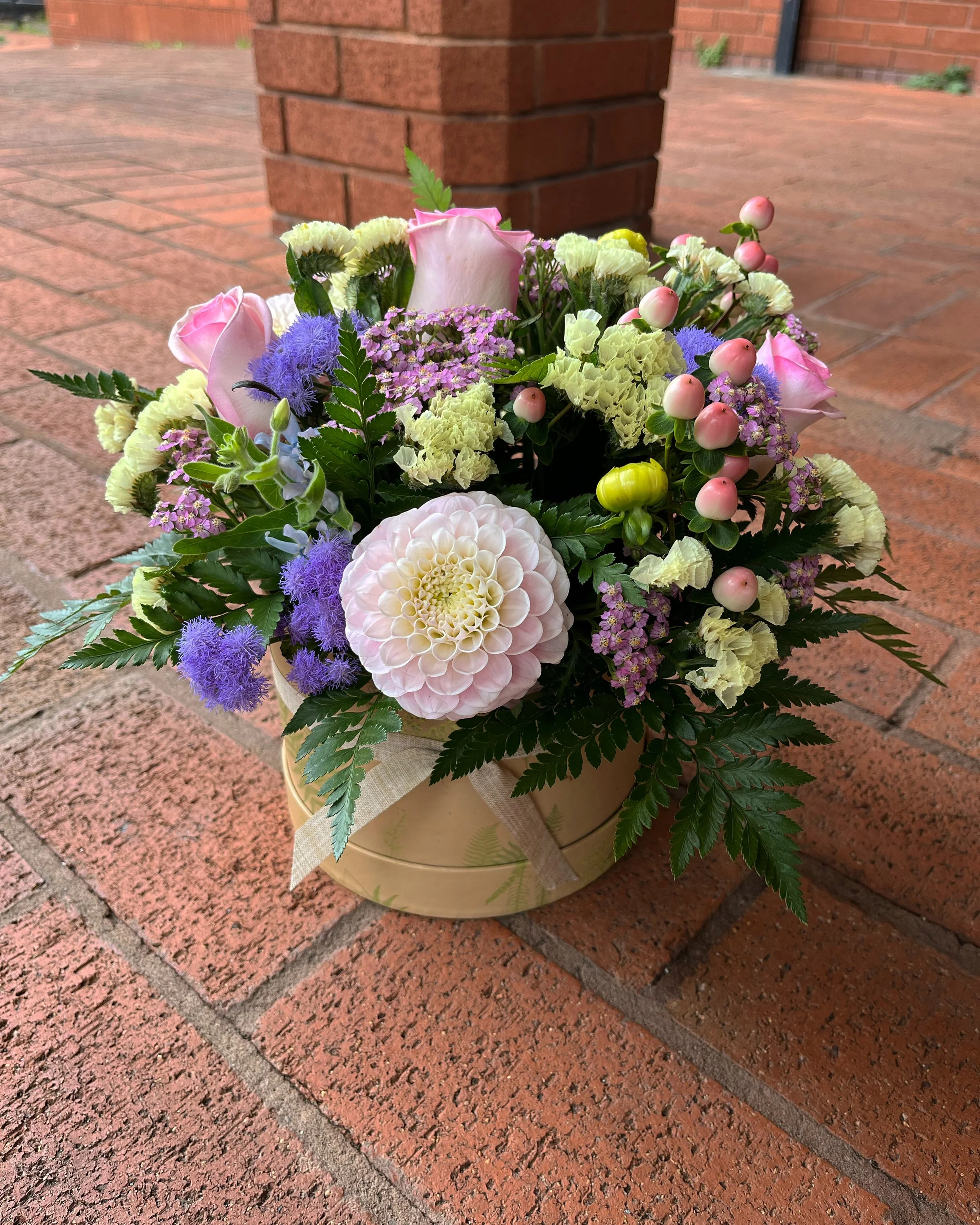 A bouquet of mixed flowers, including pink roses, white carnations, purple limonium, and other small colorful flowers, in a beige pot, placed on a brick surface with a brick wall in the background.