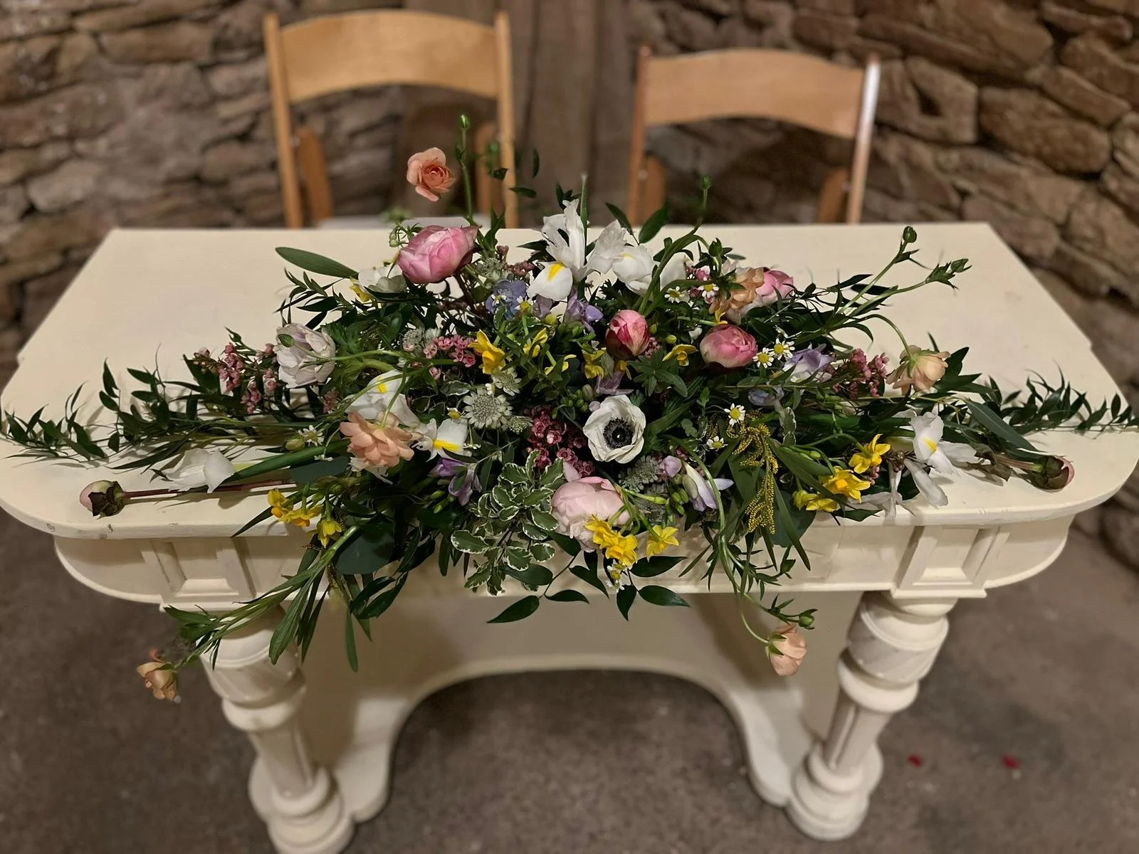 A white Grandma-style table with ornate legs, decorated with a cascading bouquet of mixed flowers including pink roses, white anemones, yellow and white small blossoms, and greenery. The table is set against a stone wall background.