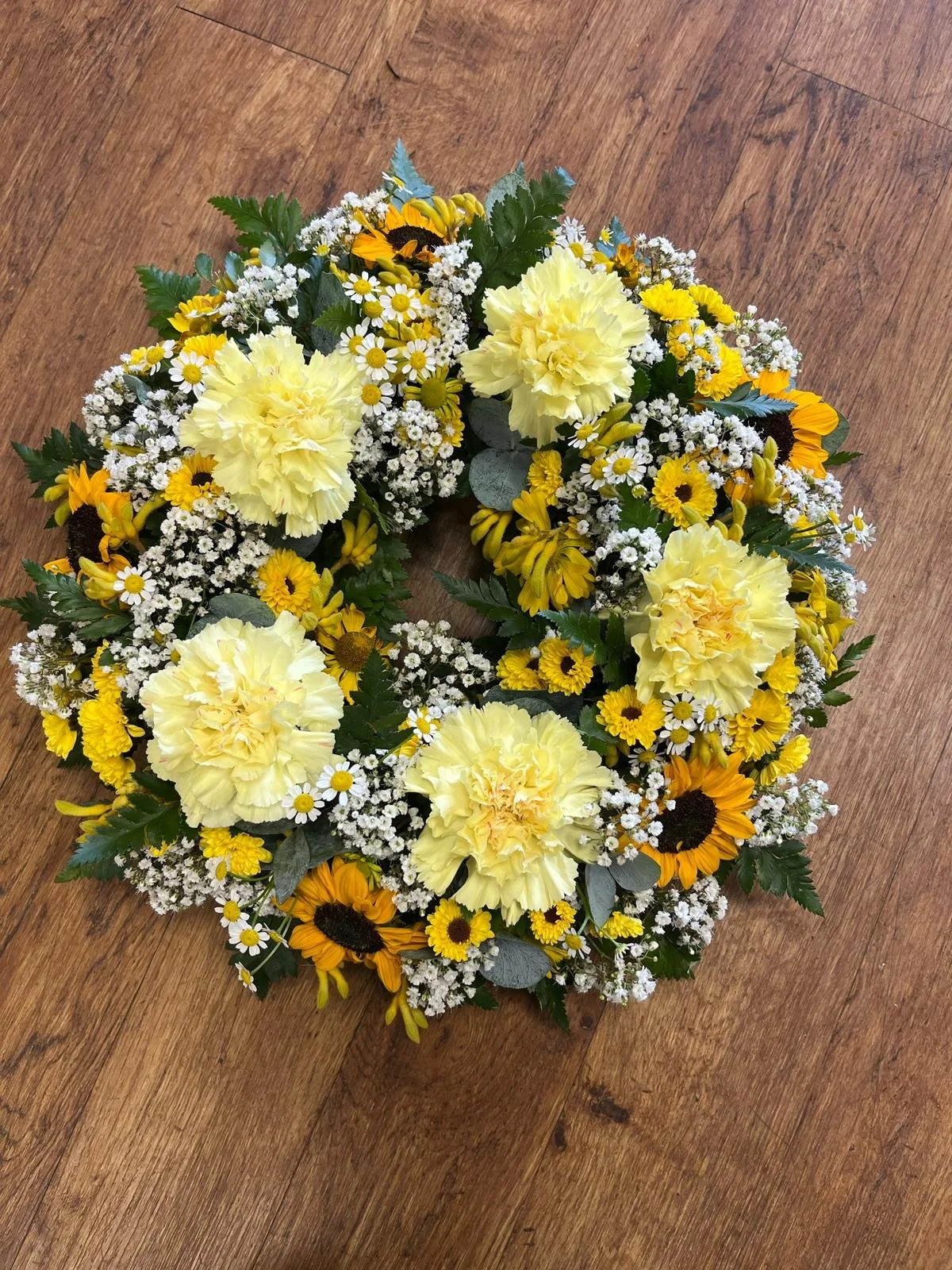 A floral wreath made of yellow carnations, daisies, chrysanthemums, baby's breath, and greenery, resting on a wooden floor.