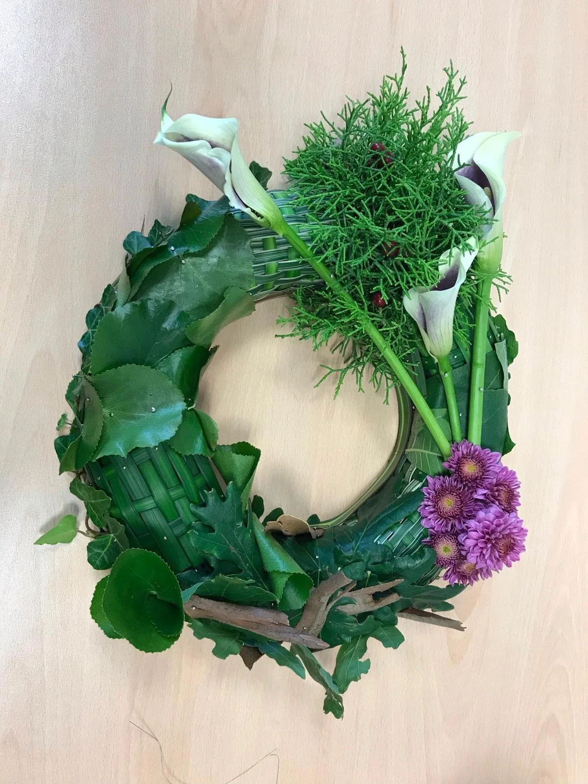 A decorative floral arrangement with calla lilies, purple daisies, green foliage, and a woven basket, arranged in a circular pattern on a beige wall.