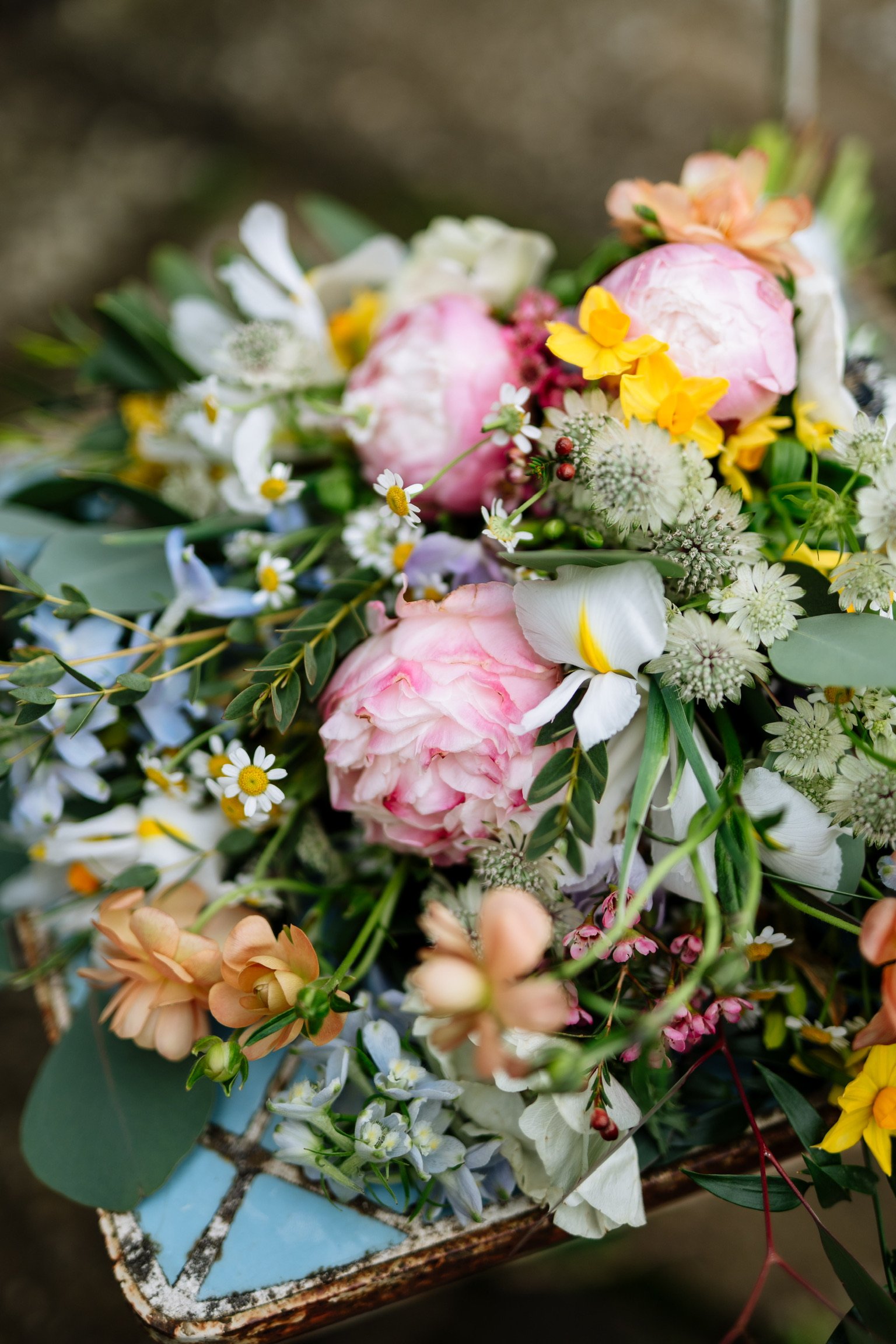 A bouquet of various flowers, including pink peonies, white daisies, yellow blooms, and greenery, resting on a rustic, light blue tray.