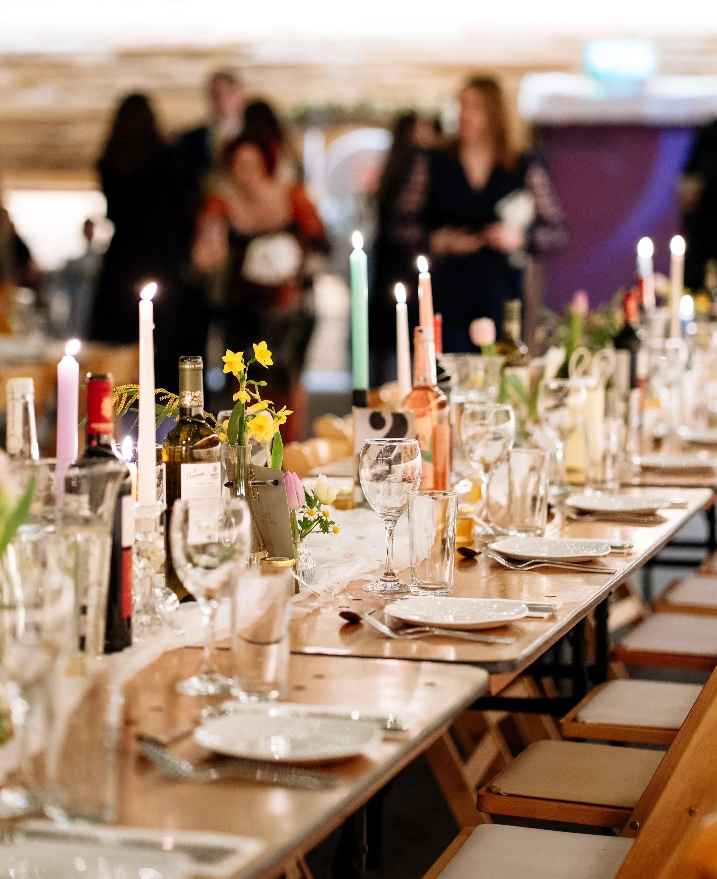 A decorated dining table with candles, flowers, wine bottles, glasses, and tableware, set for a celebration or gathering, with people in the background talking and socializing.