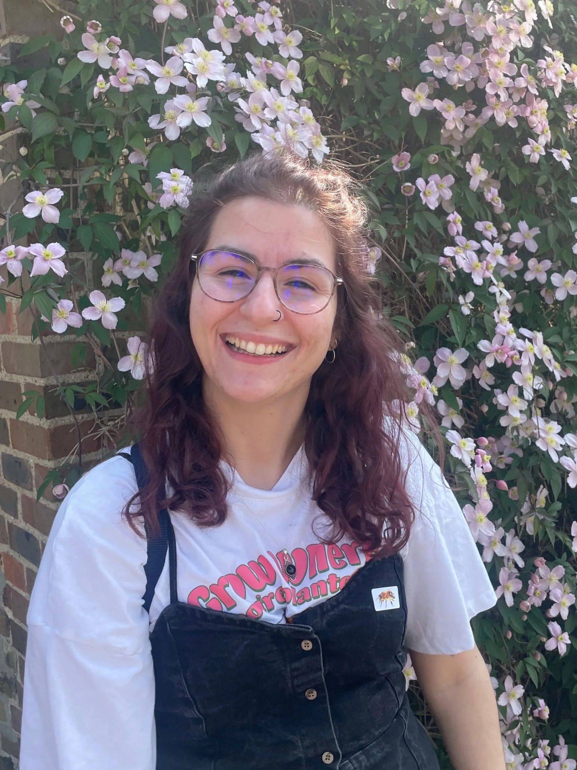 A smiling woman with curly dark red hair, wearing glasses, a nose ring, and a white t-shirt with colorful text, standing in front of a bush with pink and white flowers.
