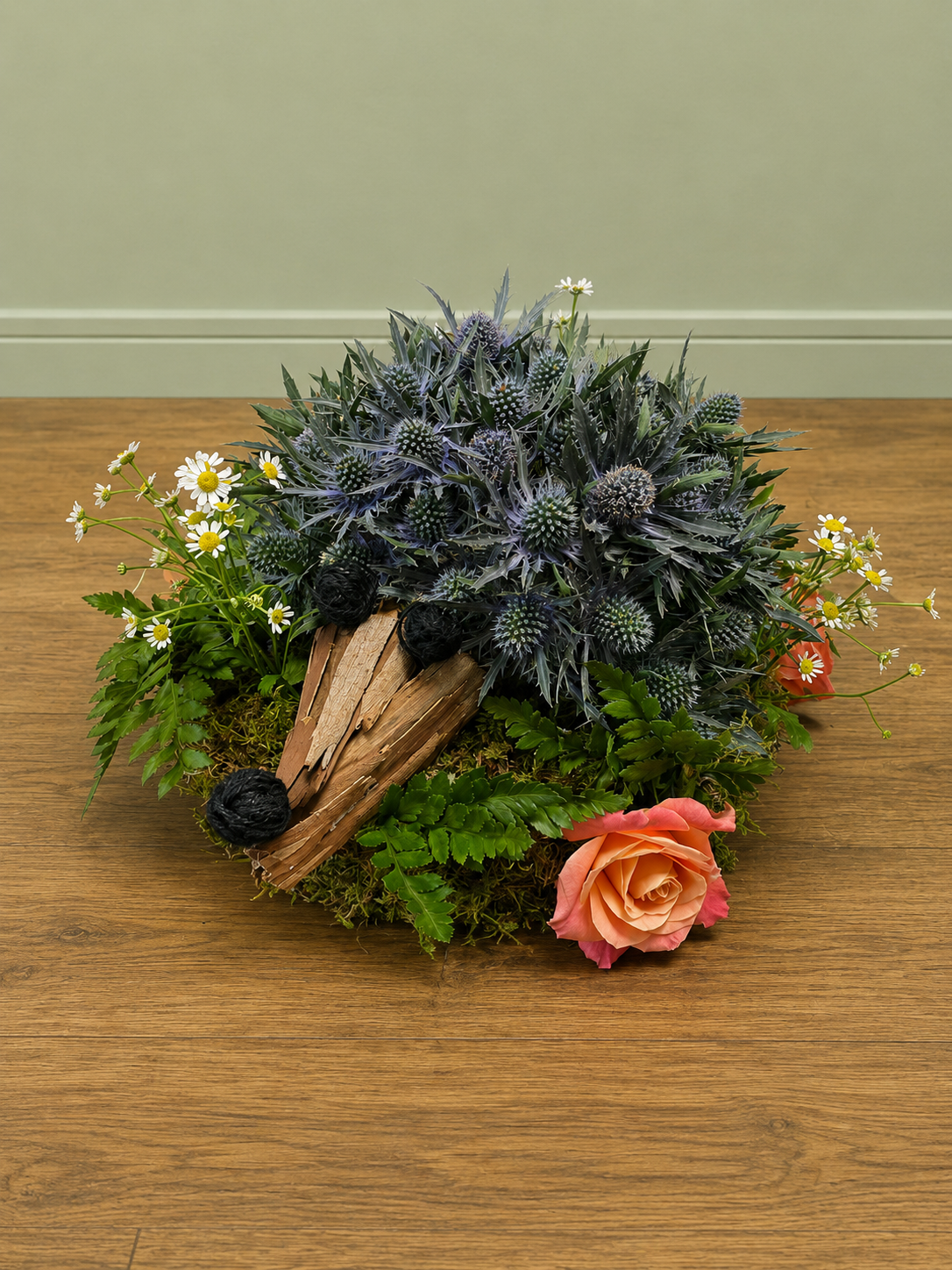 A floral arrangement on a wooden surface with purple and green spiky plants, small white daisies, a peach-colored rose, and decorative wood and moss.