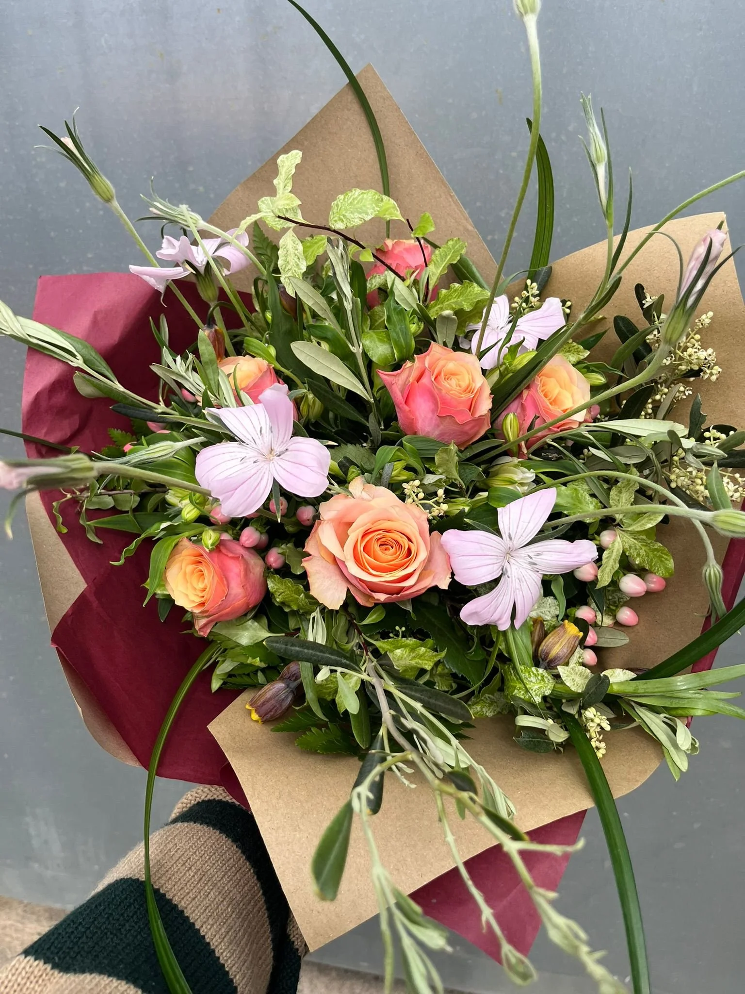 A colorful bouquet of flowers including peach roses, light pink lilies, and various greenery, wrapped in brown and red paper.