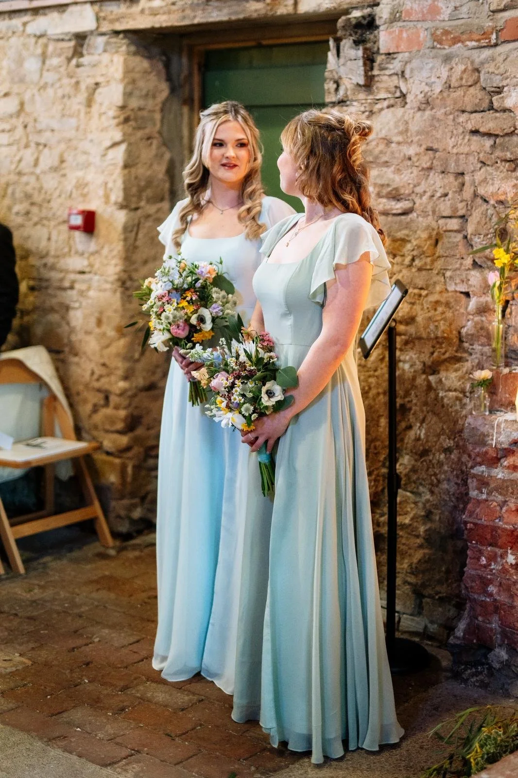 Two women in light blue dresses standing indoors with a stone wall background, holding bouquets of flowers, likely at a wedding or special event.