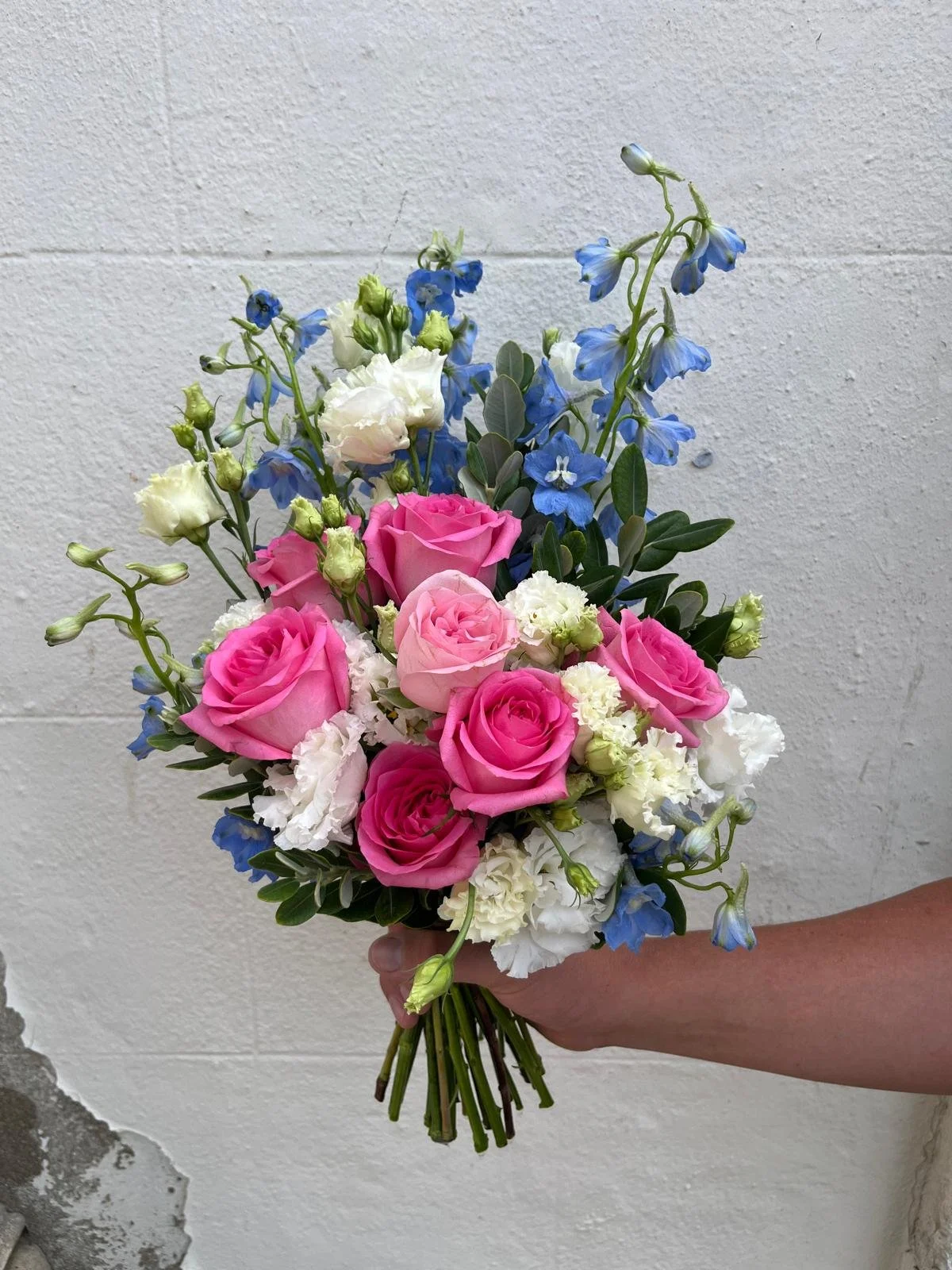 Hand holding a bouquet of pink roses, white carnations, and blue delphinium flowers against a white brick wall.