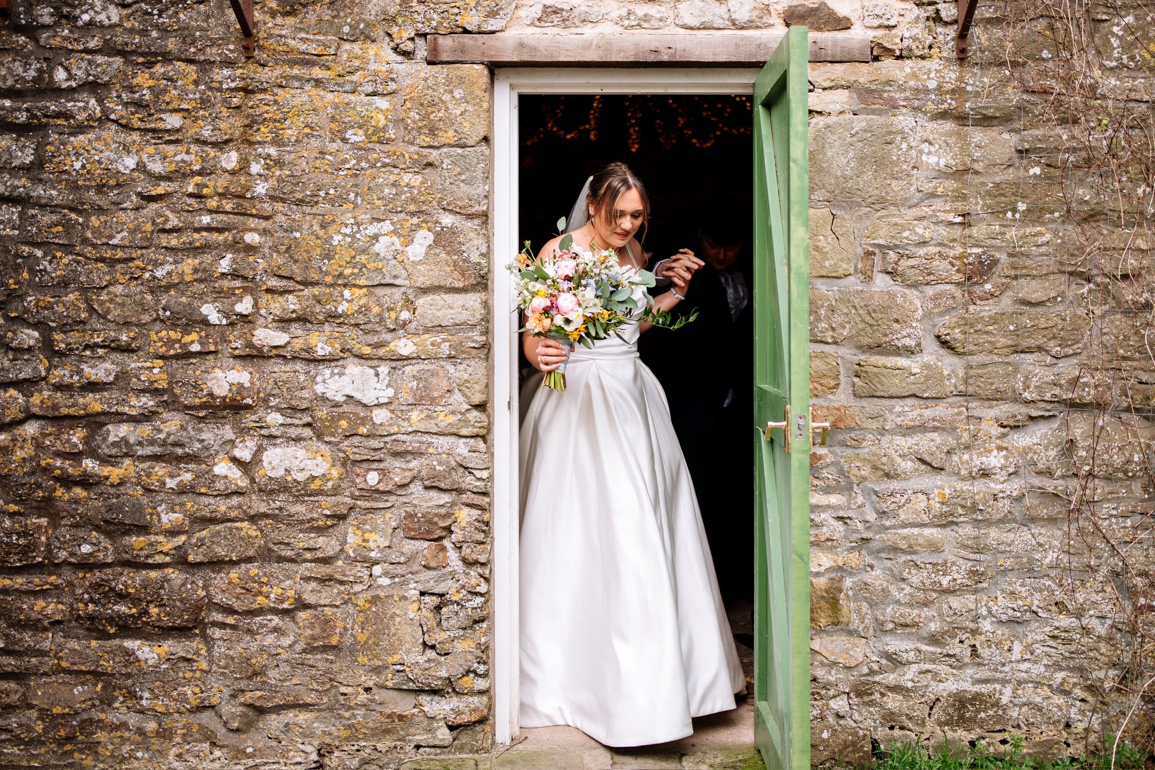 A bride in a wedding gown holding a colorful bouquet, standing in an open window of a rustic stone wall building, with a man beside her inside.