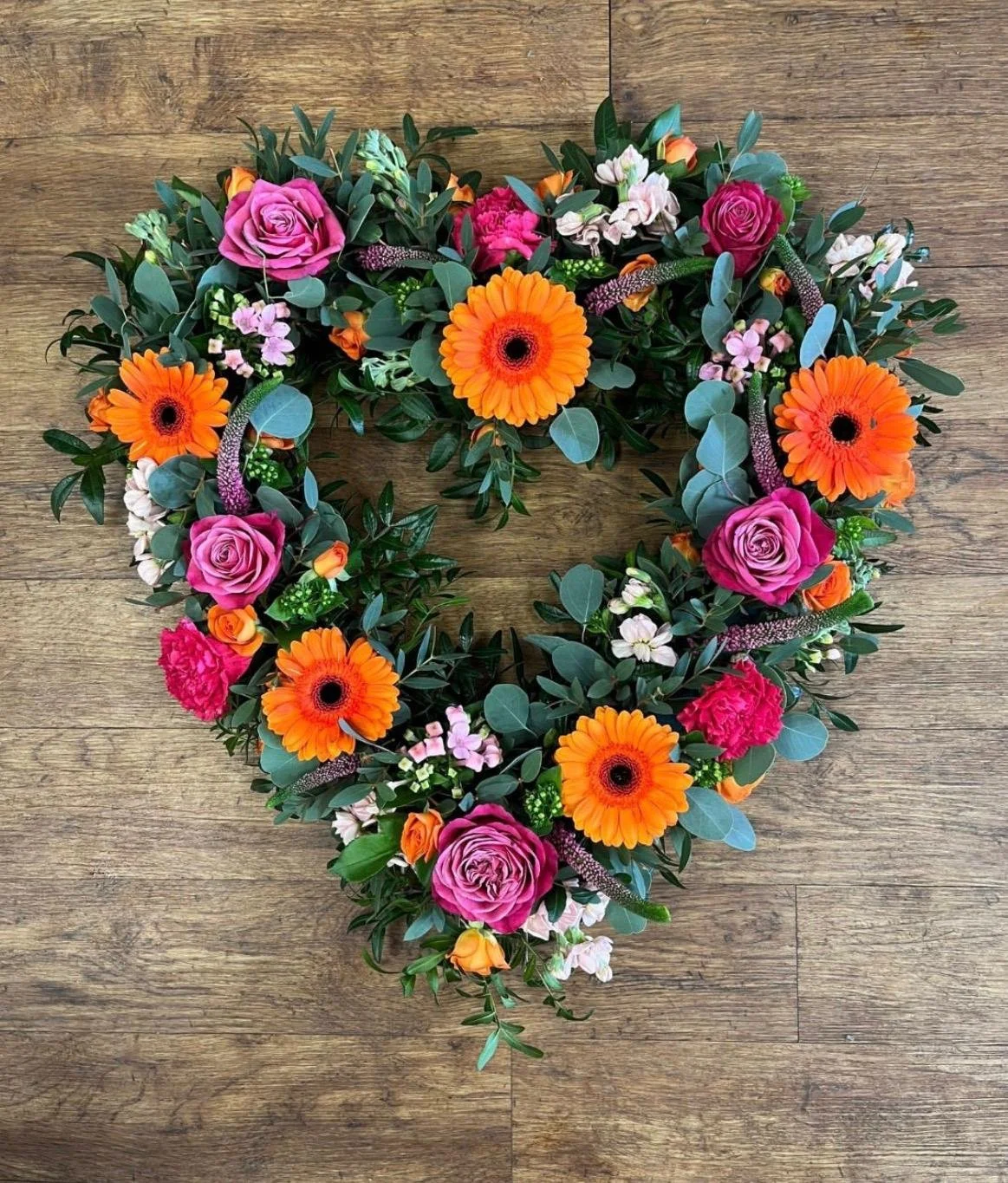 Heart-shaped floral arrangement with pink roses, orange gerbera daisies, pink carnations, and various green leaves on a wooden surface.