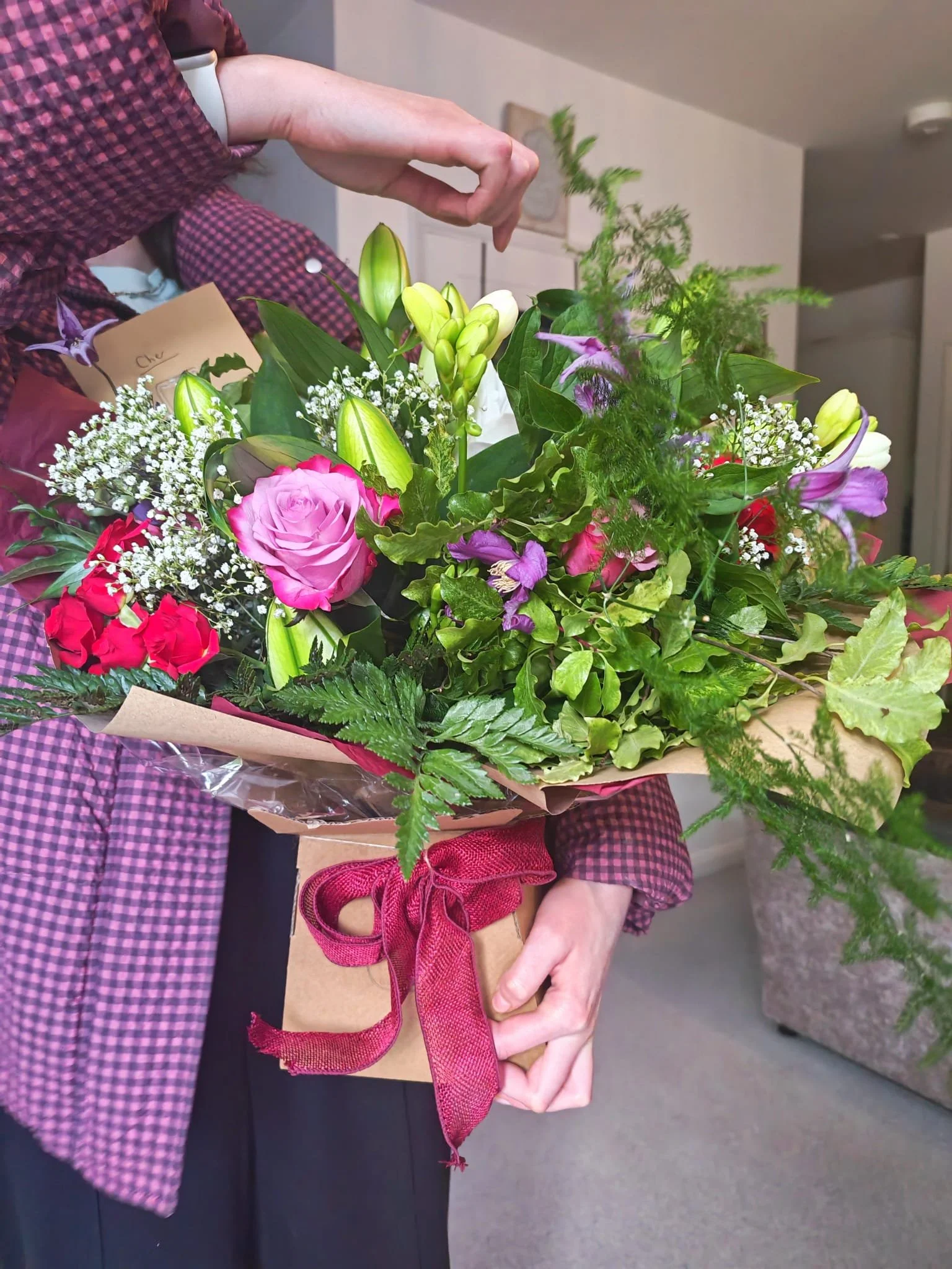 Person holding a bouquet of pink, white, purple, and green flowers wrapped in brown paper with a red ribbon.
