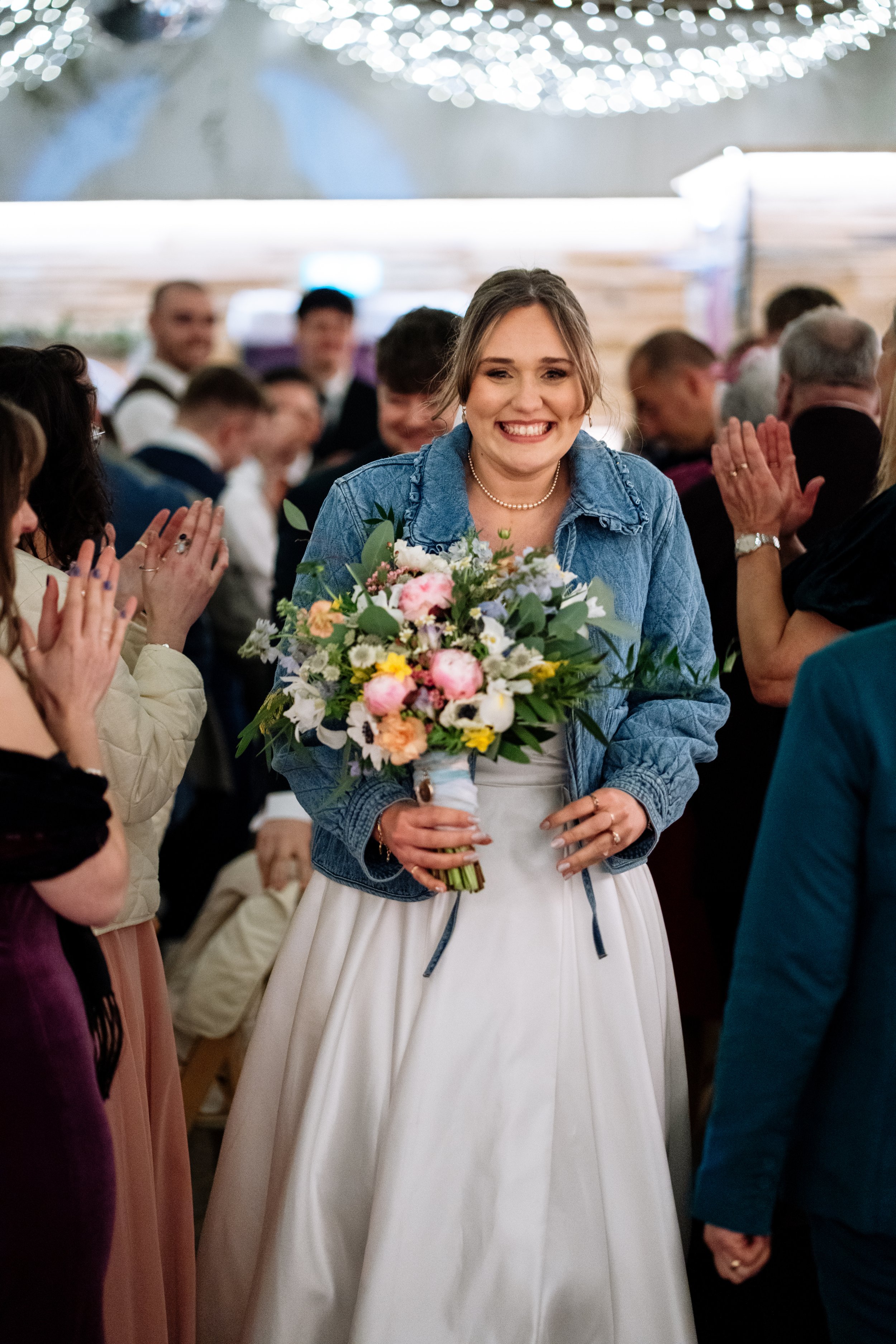 A woman in a white dress and denim jacket holding a large bouquet of pink, white, and purple flowers, smiling at a celebration or wedding. She is surrounded by people clapping and celebrating in an indoor venue decorated with lights.