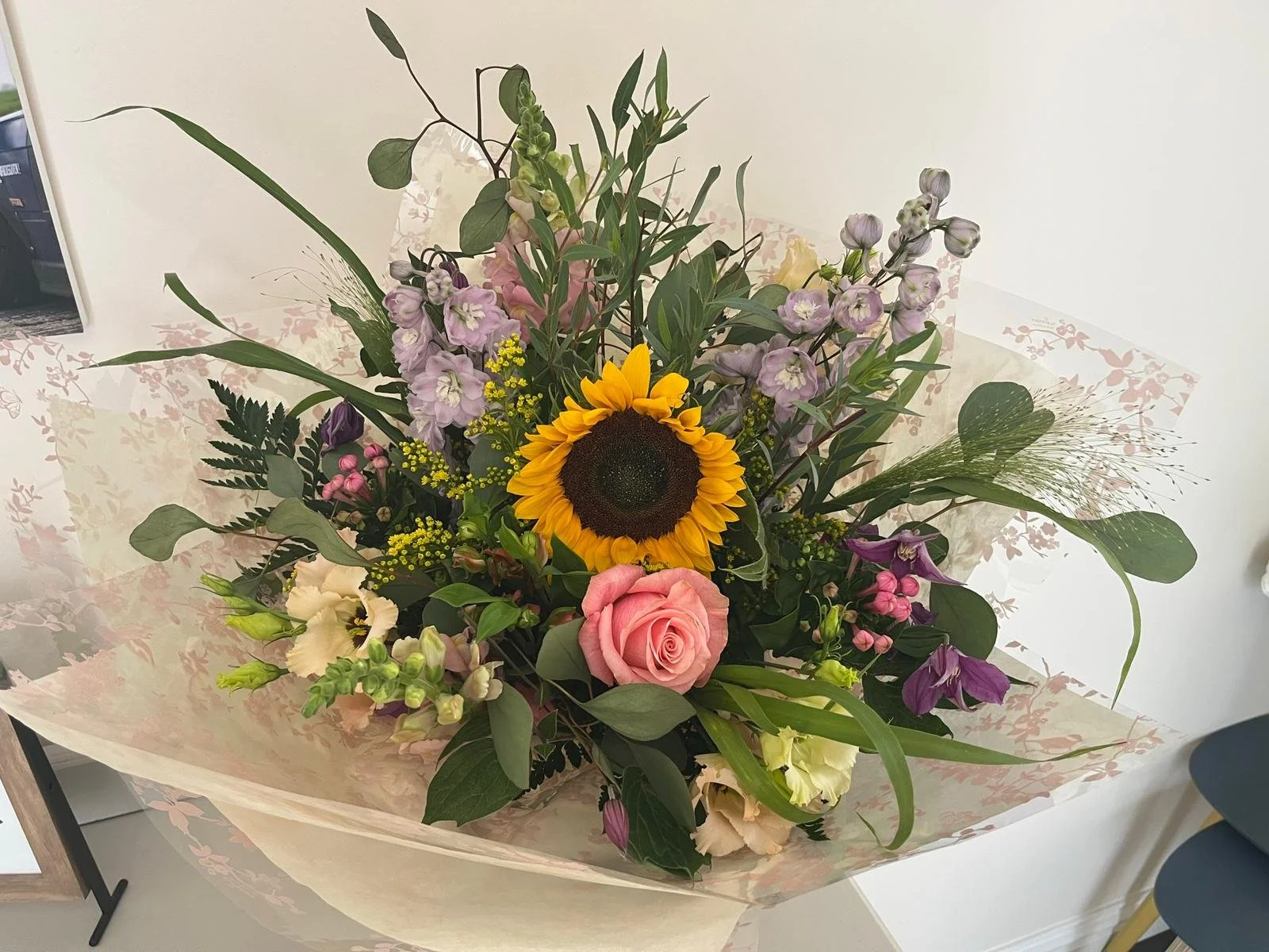 A colorful flower bouquet with a sunflower, pink rose, purple flowers, yellow accents, green leaves, and filler flowers wrapped in floral paper.